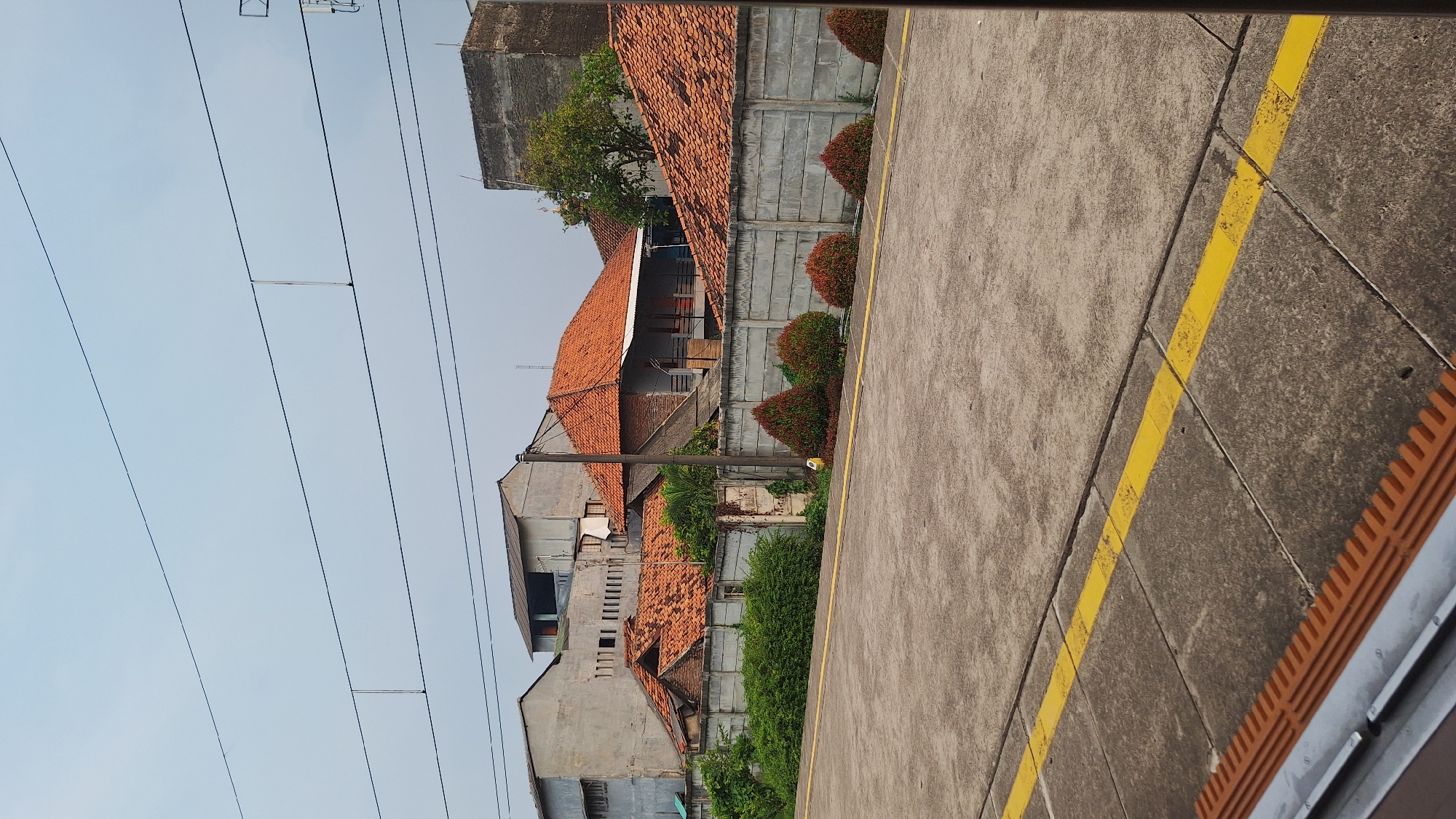 The image depicts a street scene in Cikarang, Indonesia. The view is taken from an elevated position, possibly a platform or bridge. Houses with orange-tiled roofs are visible behind a concrete wall with bushes. Overhead power lines run parallel to the street. The street is marked with a yellow safety line.
