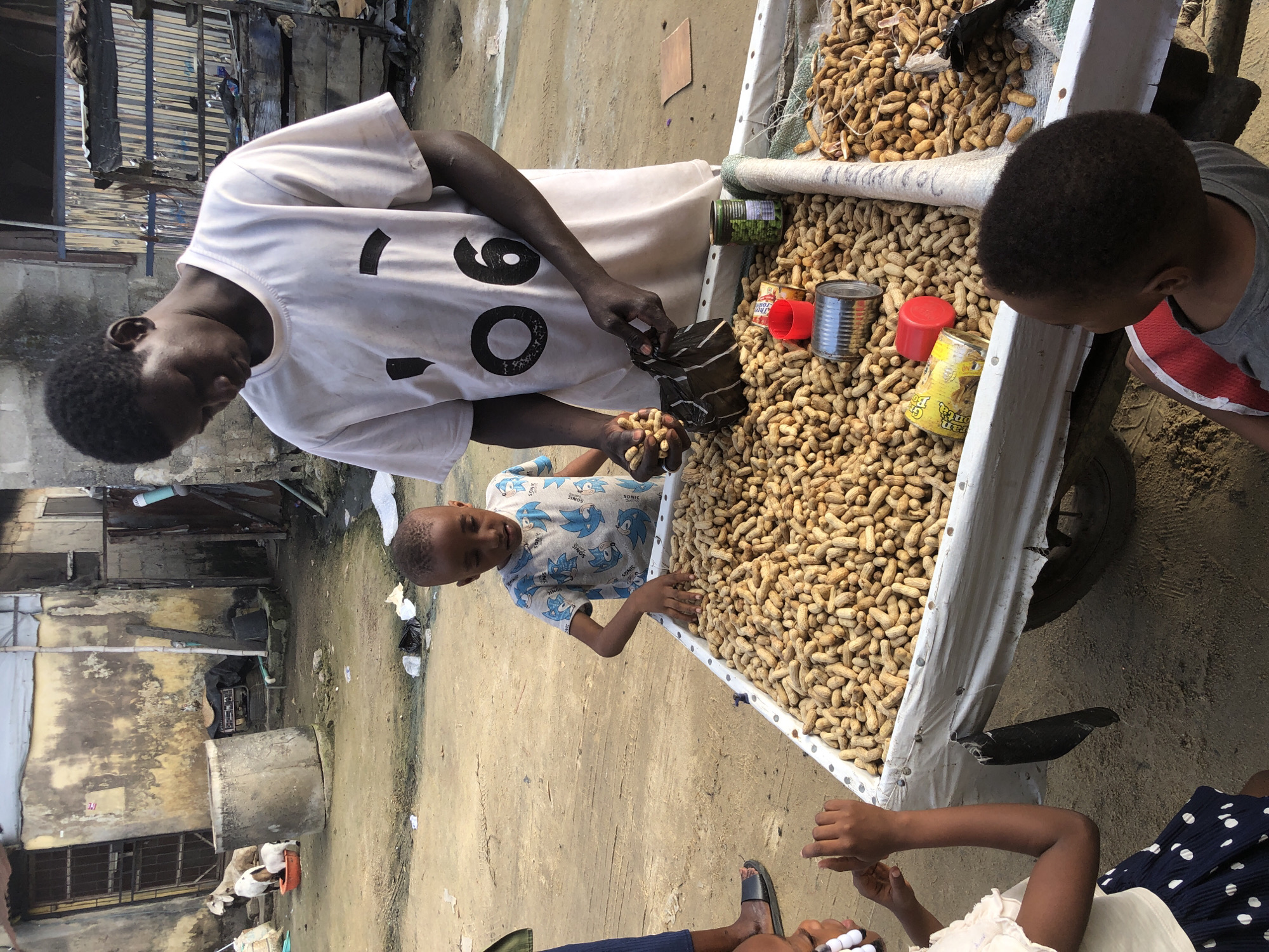 In Ado, Nigeria, a Black male vendor, wearing a light-colored t-shirt with a large graphic design, is engaged in a transaction at a peanut stand. He is holding a bag filled with peanuts and a handful of peanuts, appearing to be selling them to two young Black children. One child, a boy wearing a blue t-shirt with a character print, stands on the opposite side of the cart, reaching towards the peanuts. A younger child, a girl with dark skin and wearing a light-colored top and a blue skirt with white polka dots, is positioned to the left of the cart, looking towards the vendor. The cart itself is laden with a large quantity of peanuts, displayed in a white trough. Several cans, including a red one and a green one, are visible on top of the peanuts, as well as a small red cup. The background depicts a rustic, urban environment with weathered buildings, some appearing to be constructed of concrete blocks and corrugated metal. A concrete cylindrical structure and two goats are visible to the left of the frame, suggesting a semi-rural or informal urban setting. The ground is unpaved, with visible tire tracks.