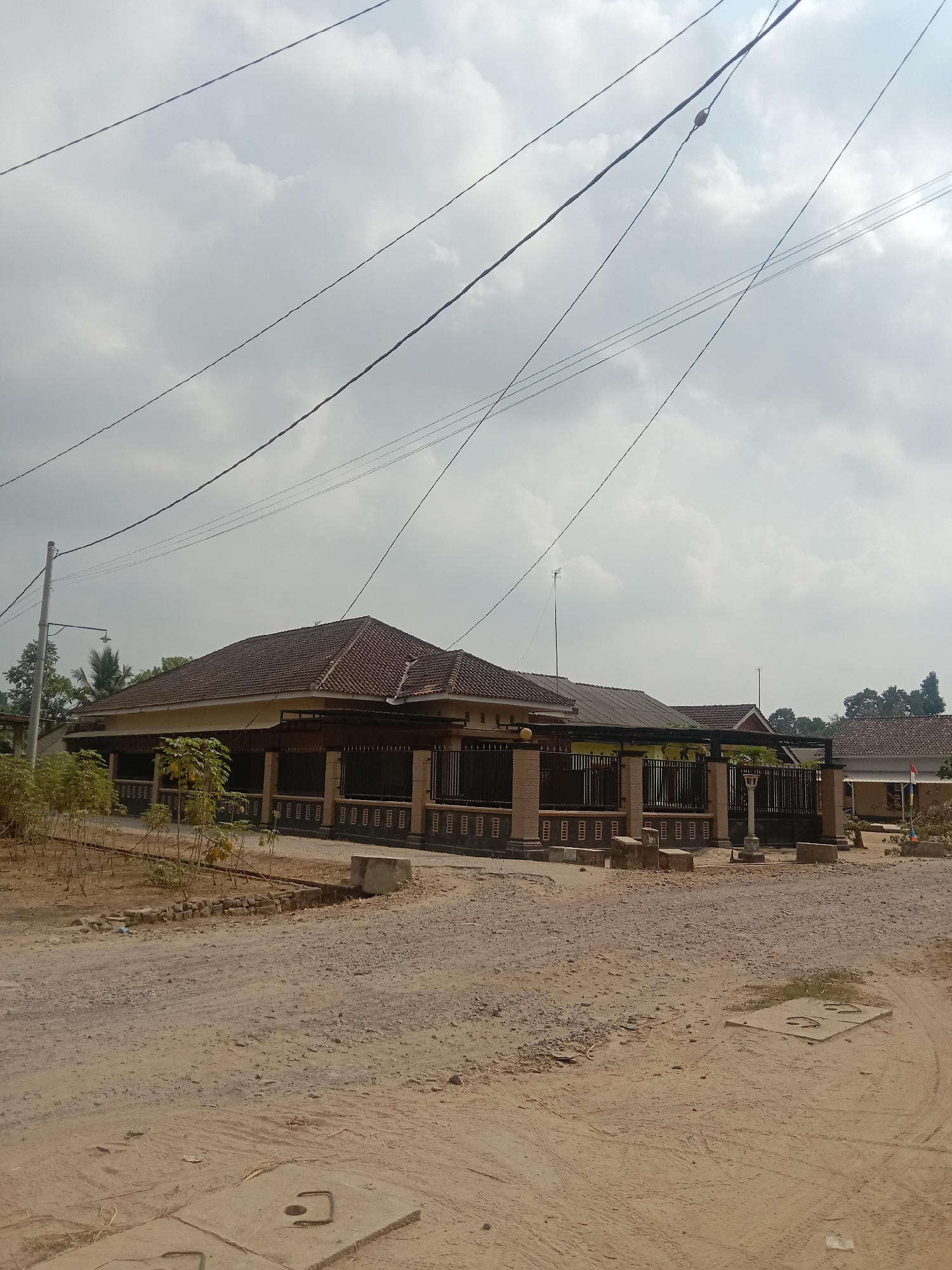 The image depicts a street scene in Utama Jaya Mataram, Indonesia, characterized by a dirt road leading towards a residential property. A low-lying house with a tiled roof is visible, bordered by a fence with decorative columns. Several power lines crisscross the sky, which is partly cloudy. Sparse vegetation, including small trees and bushes, dots the foreground and the area around the house. The ground is a mix of dirt and gravel, with concrete slabs, possibly manhole covers, present on the road surface. The overall impression is that of a semi-rural or developing area. No people are visible in the frame.