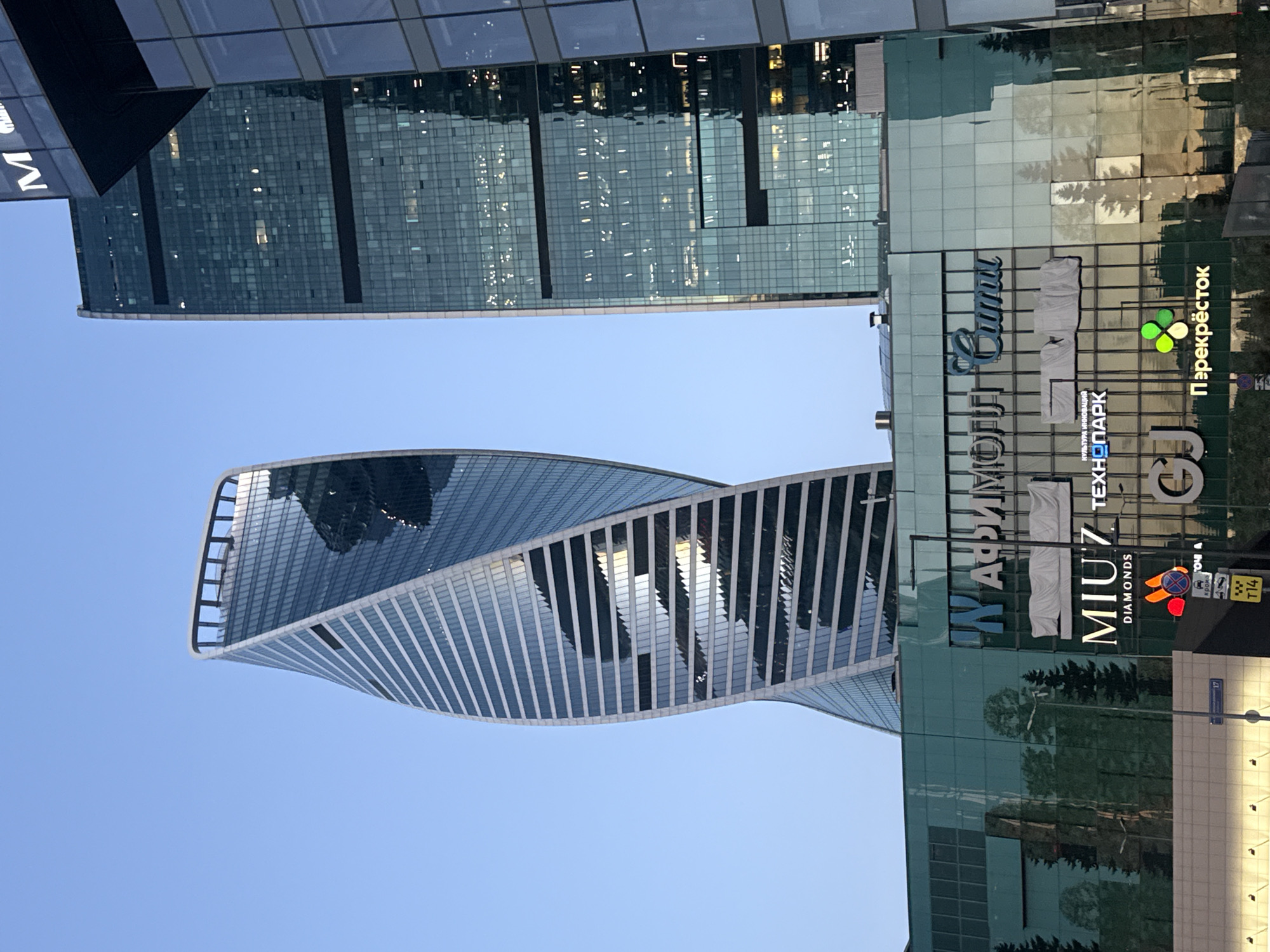The image shows a view of modern skyscrapers in Khimki, Russia, under a clear blue sky. In the foreground, a multi-story building with a glass facade reflects trees and the sky. Several signs are visible on this building, including "MIU'7 DIAMONDS", "ТЕХНОПАРК", and "Перекрёсток". To the left and center, a distinctive twisted skyscraper dominates the view, its glass exterior reflecting the surrounding buildings and sky. To the right, another tall, rectangular skyscraper with a green glass facade rises, its windows illuminated. The scene appears to be captured during daylight, possibly late afternoon given the warm reflections on the glass. There are no people clearly visible in the image.