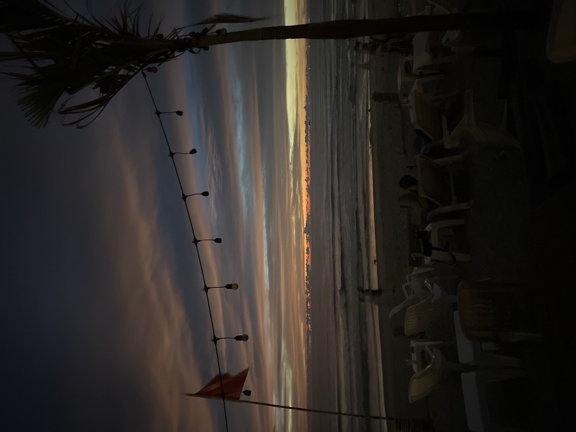 The image depicts a beach scene at dusk. A string of lights hangs across the frame, with a few bulbs visible. A red flag is planted on a pole on the left side. The ocean stretches out to the horizon, with waves gently lapping at the shore. The sky is filled with dramatic clouds, displaying hues of orange, pink, and blue, indicating the setting sun. In the foreground, several beach chairs and tables are scattered on the sand, some occupied by silhouetted figures. A palm tree is visible on the right, its fronds dark against the twilight sky. The overall atmosphere is serene and twilight.