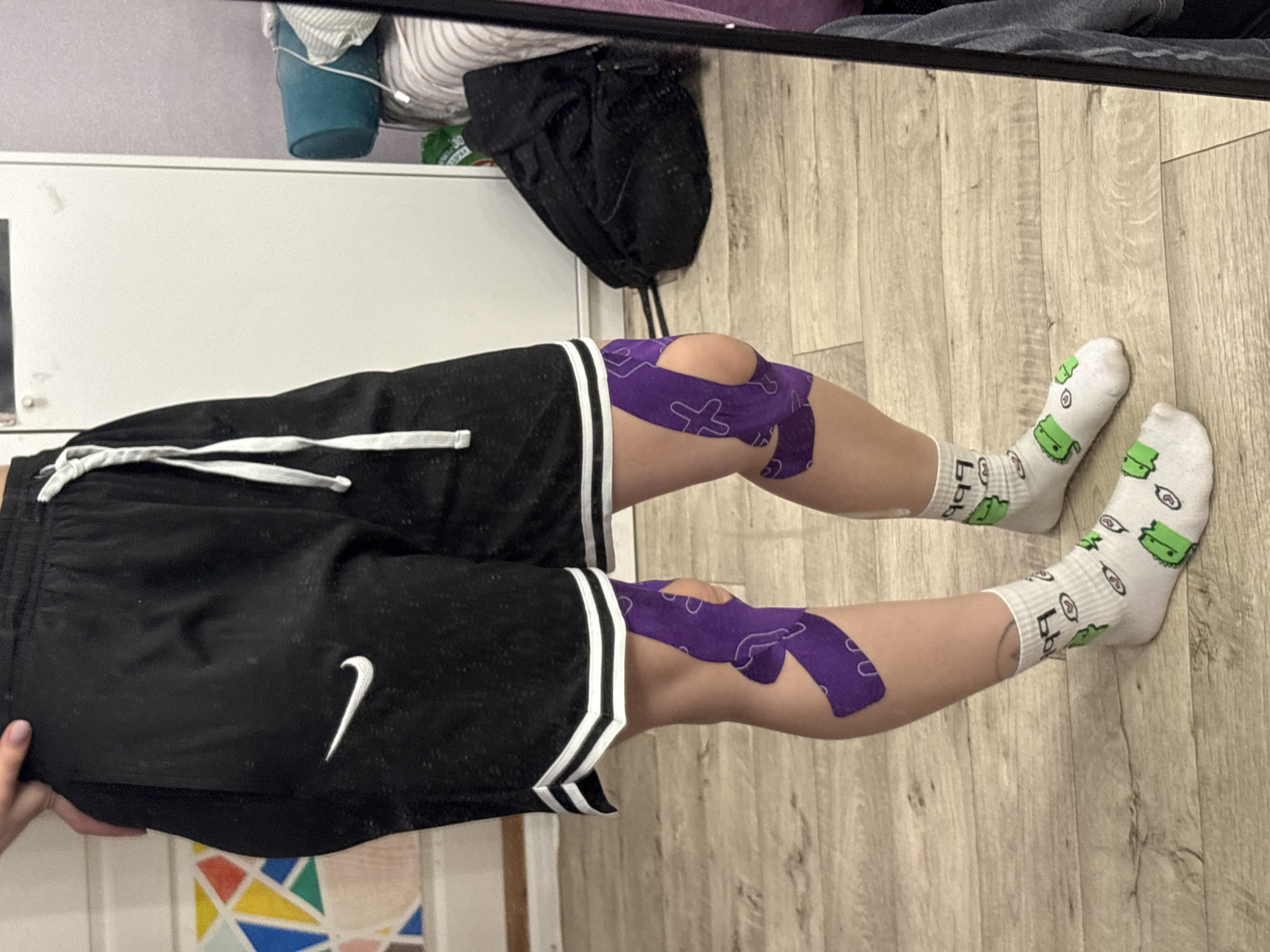 The image shows a person's legs wearing black athletic shorts with a white Nike logo and white trim. Both knees are covered with purple kinesiology tape. The person is wearing white socks with green cartoon designs. The background includes a wooden floor, a white cabinet with items on top, and a reflection of an object above the cabinet. There is no information to associate this photo with Прикамский, Russia.