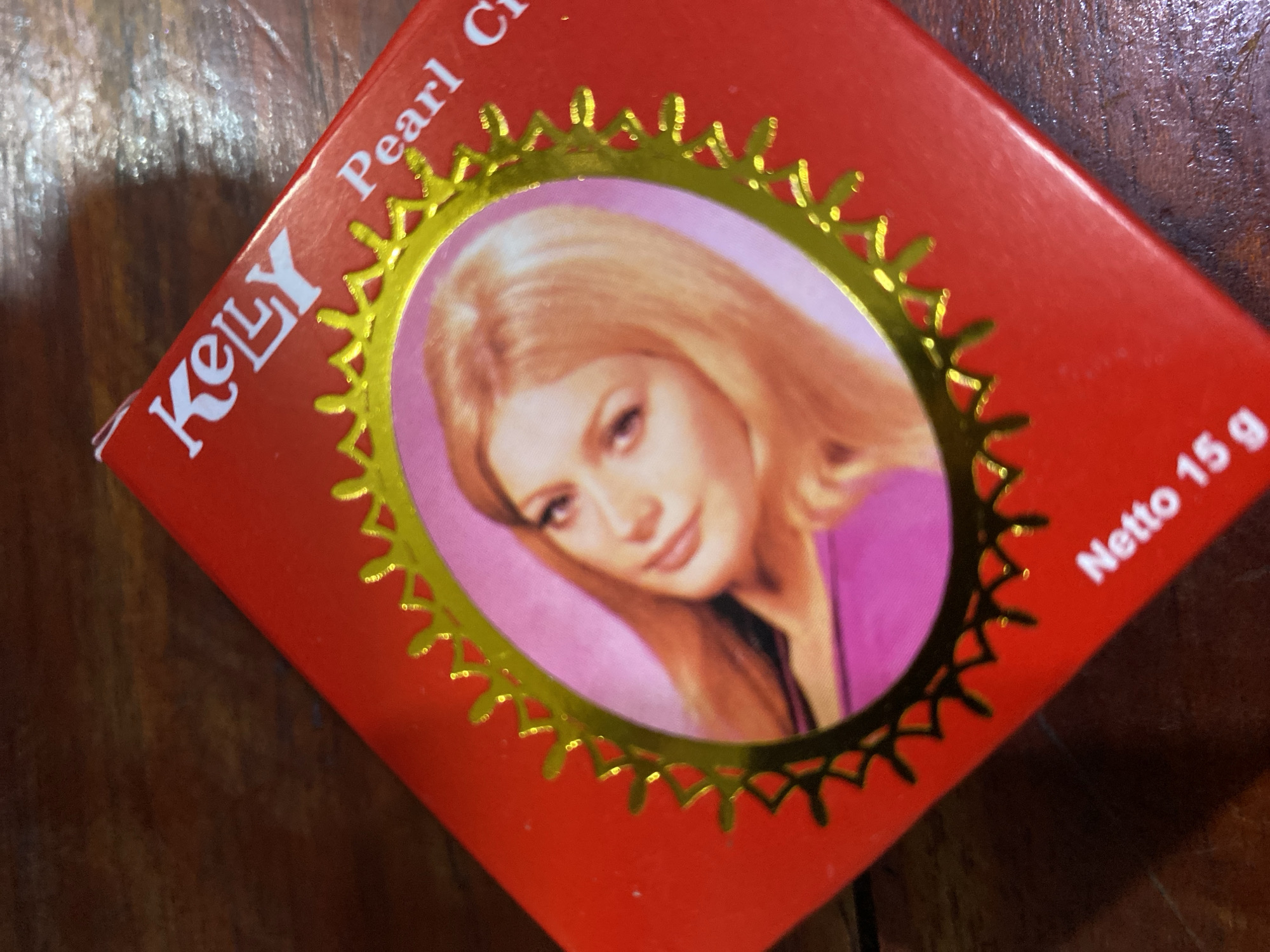 The image shows a close-up of a red box with a gold decorative border. Inside the border is a picture of a blonde woman's face. The words "KELLY Pearl Cr" are visible in white text on the box. Below the image, the words "Netto 15 g" are visible in white text. The box is placed on a wooden surface. There is no indication of a city or specific location in the image.