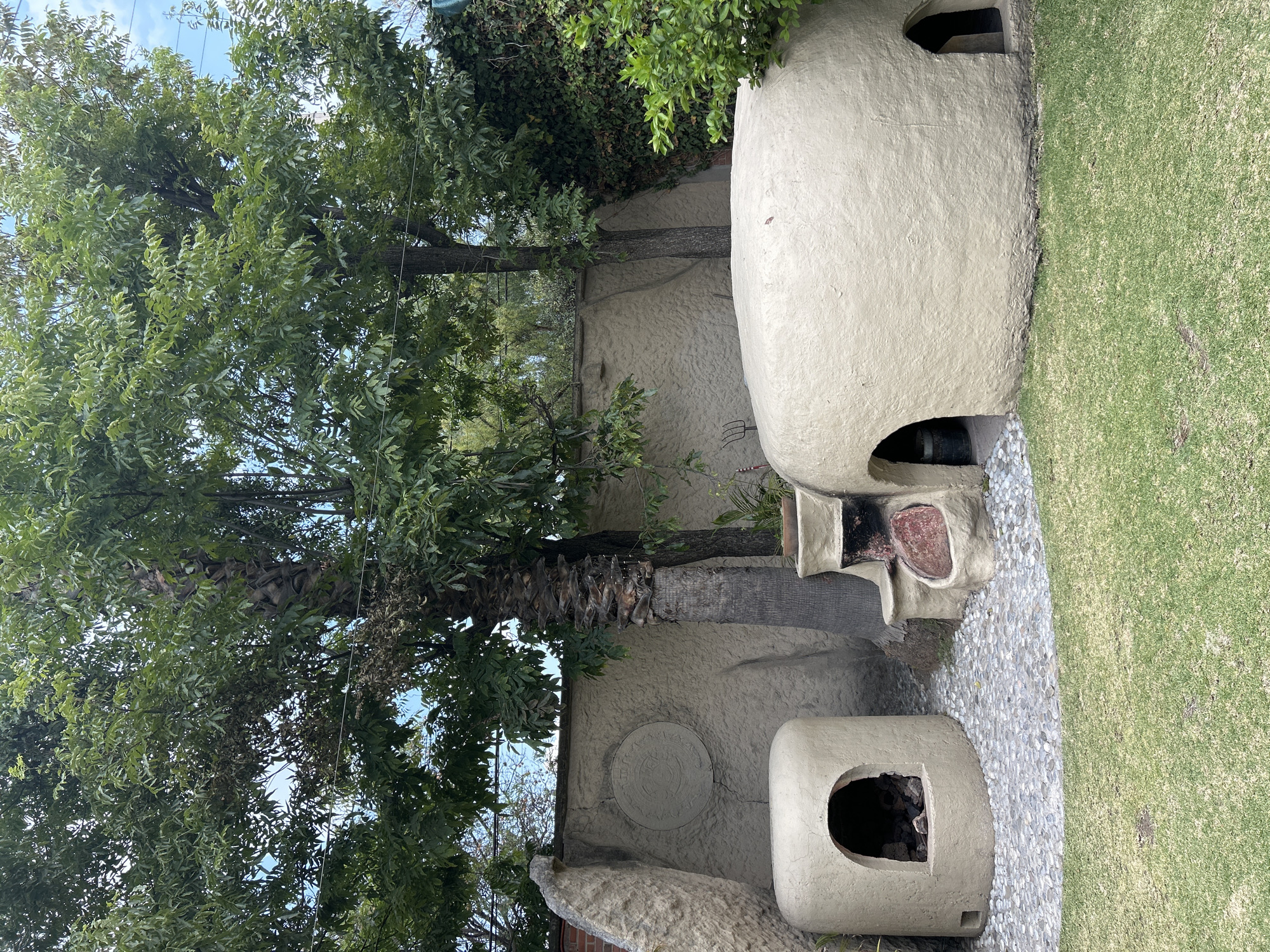The image shows an outdoor scene in Unknown, Unknown, featuring architectural elements and greenery. A large, beige, domed structure, possibly an outdoor oven or architectural feature, dominates the right side of the frame. Adjacent to it is an area covered with small, light-colored pebbles, leading to a patch of well-maintained green grass.
To the left, a mature tree with dense, green foliage fills the frame, partially obscuring a wall with a textured, light-colored surface. A round, decorative element is affixed to the wall. Below the tree is a smaller, similarly shaped structure to the large dome, also with an opening, suggesting a functional or decorative purpose. The general environment is reminiscent of a backyard or garden setting.