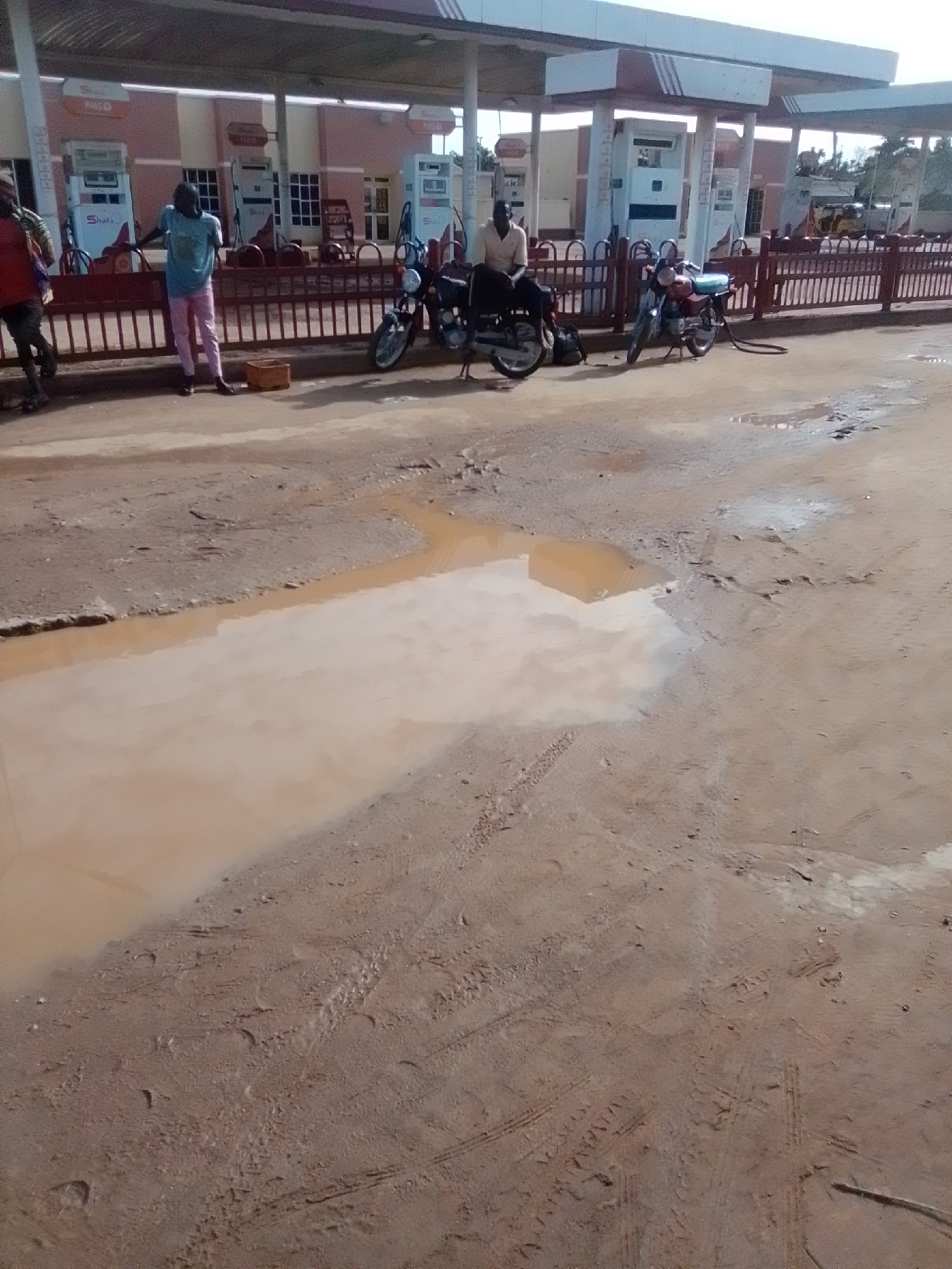 The image depicts an outdoor scene at a gas station in Kwadon, Nigeria. A portion of the gas station building with several fuel pumps is visible in the background. The ground in the foreground is a dirt road with a large puddle of muddy water. Three individuals are present, two of whom are standing and one is sitting on a motorcycle. Two motorcycles are parked near the gas station. The overall condition of the road and the presence of the puddle suggest recent rainfall.