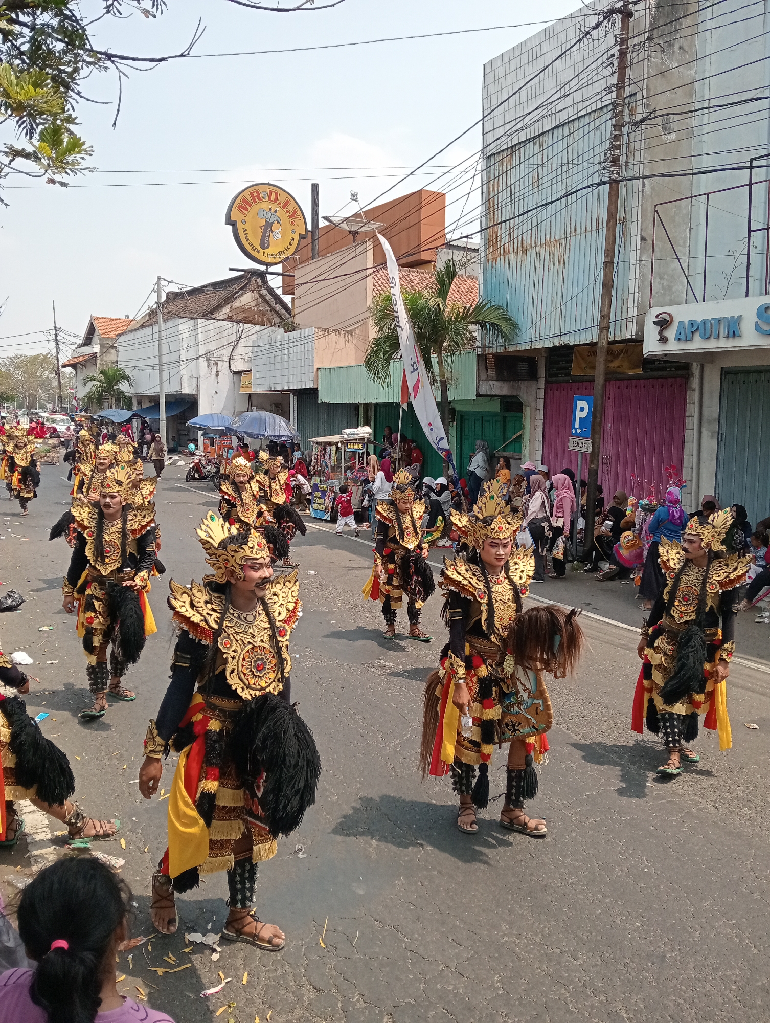 The image depicts a street parade in Demak, Indonesia, where several individuals are dressed in elaborate, traditional golden and black costumes with feathered headdresses and tribal markings. They appear to be performers, possibly participating in a cultural festival or celebration. Some are carrying faux horse heads, suggesting a dance or theatrical representation. They are marching down a paved street lined with shops and buildings. Spectators are visible on the sidewalks, observing the procession. The sky is clear and the weather appears to be sunny and warm. The shops and buildings suggest a commercial or residential area within the city.
