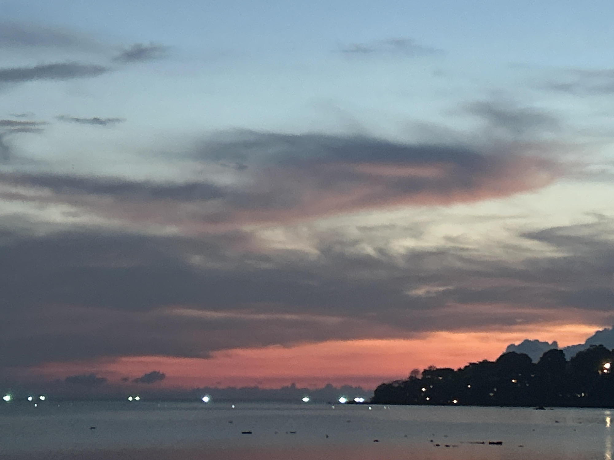 The image captures a twilight scene in Baan Sri Thanu, Thailand. In the foreground, a body of water stretches out with small dark shapes floating on its surface. Along the distant shoreline, a dark land mass is defined by trees. At intervals along the coast, lights are visible, some reflected in the water. The sky is filled with clouds, their colors varying from pink and red near the horizon to gray and blue further up.