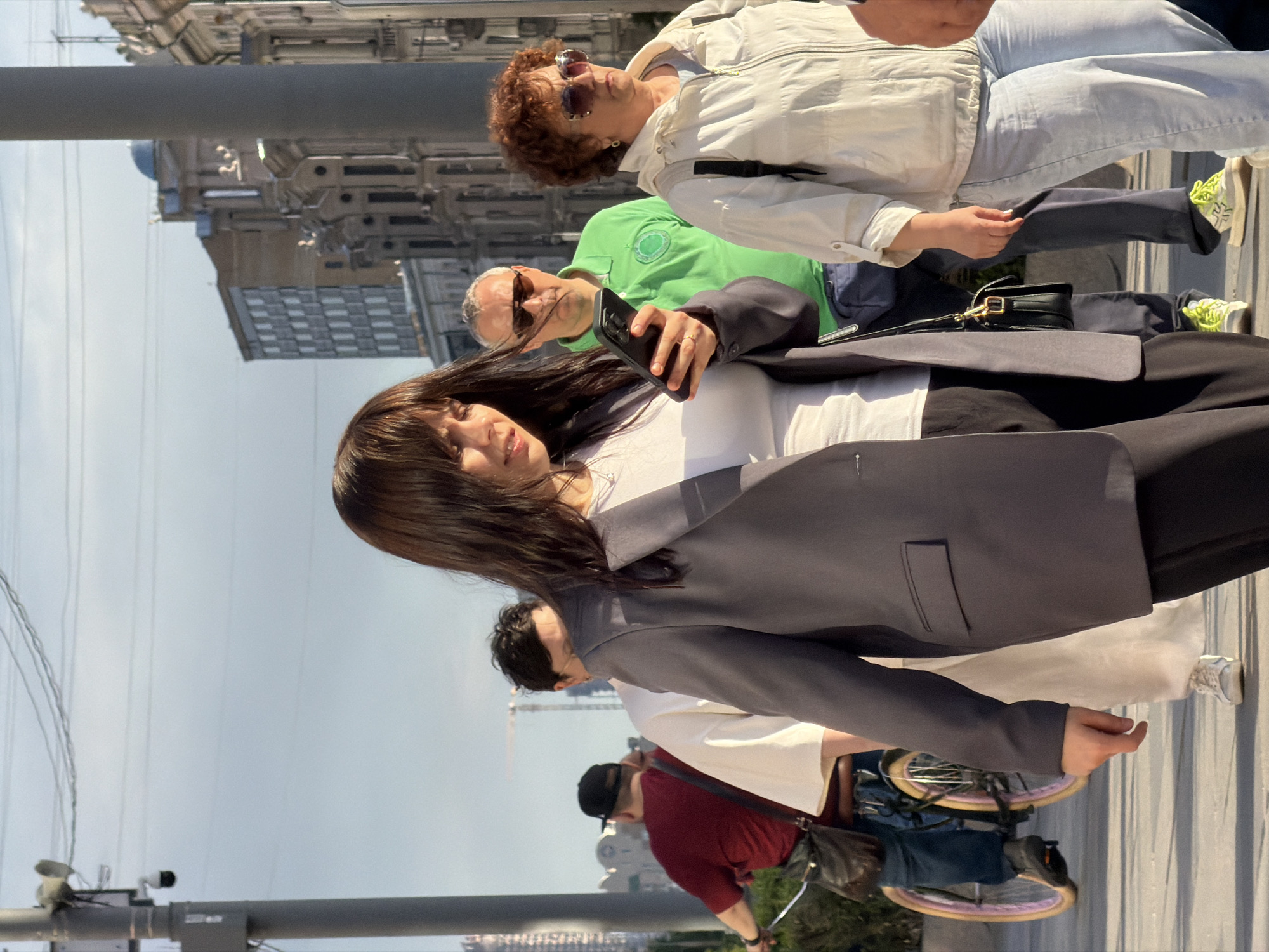 The image depicts a group of people walking on a city street in an urban setting. The main subject is a woman in a white top and black pants, with a grey blazer draped over her shoulders. She is holding a smartphone. Behind her, a man in a green shirt appears to be using a phone as well. An older woman in a white jacket and light blue jeans walks alongside them.
In the background, partially obscured, are people walking on a sidewalk. One individual is wearing a red shirt, a black cap, and is accompanied by a bicycle with a blue bag attached. Tall buildings are visible in the distance. There are also lampposts and cables overhead, typical of a city environment. The scene seems to be captured during daytime with good lighting.