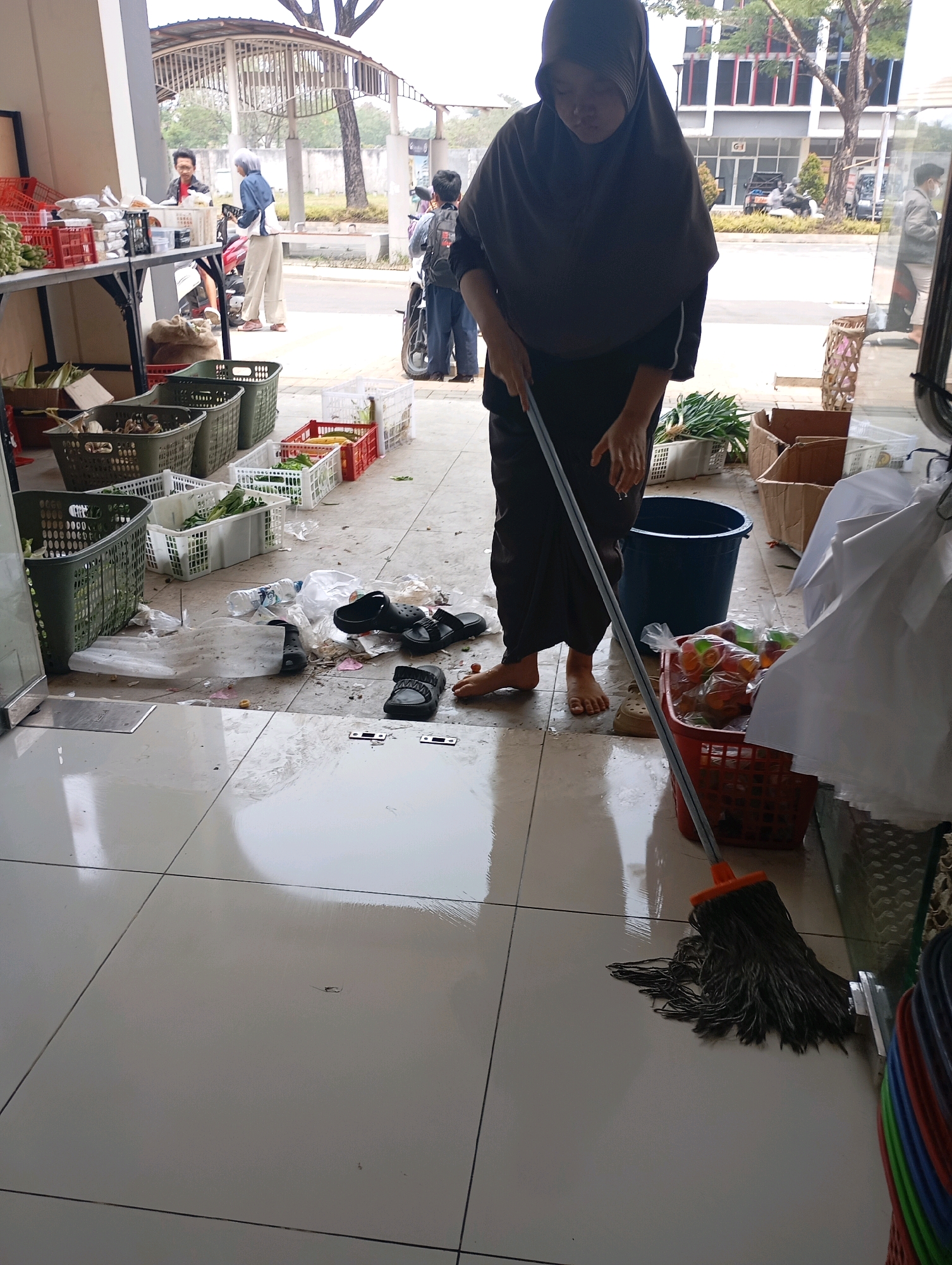 In Ciakar, Indonesia, a person wearing a dark hijab and a long, dark brown skirt is mopping a wet, tiled floor. The mop, with a dark gray string head and orange accents, is being used to clean scattered debris. Among the debris are plastic bags, food waste, and two pairs of black sandals. To the left of the mopper, a dark blue bucket is visible, and to the right, a red basket holds colorful items, possibly food. In the background, several plastic crates containing vegetables are arranged on the floor, and some people are visible in the distance, interacting near a stall or shop. The environment appears to be an outdoor or semi-outdoor area, possibly a market or a commercial establishment, with trees and buildings in the background.