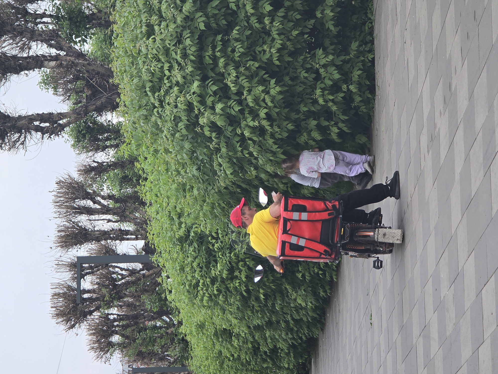 A person wearing a yellow shirt, black pants, and a red cap rides a motorcycle with a large red delivery box on the back, moving along a paved sidewalk lined with a dense green hedge. Two children stand facing the hedge, partially obscured by the foliage. In the background, trees with bare branches are visible against an overcast sky. The scene is set on a city sidewalk with a patterned paving stone surface.