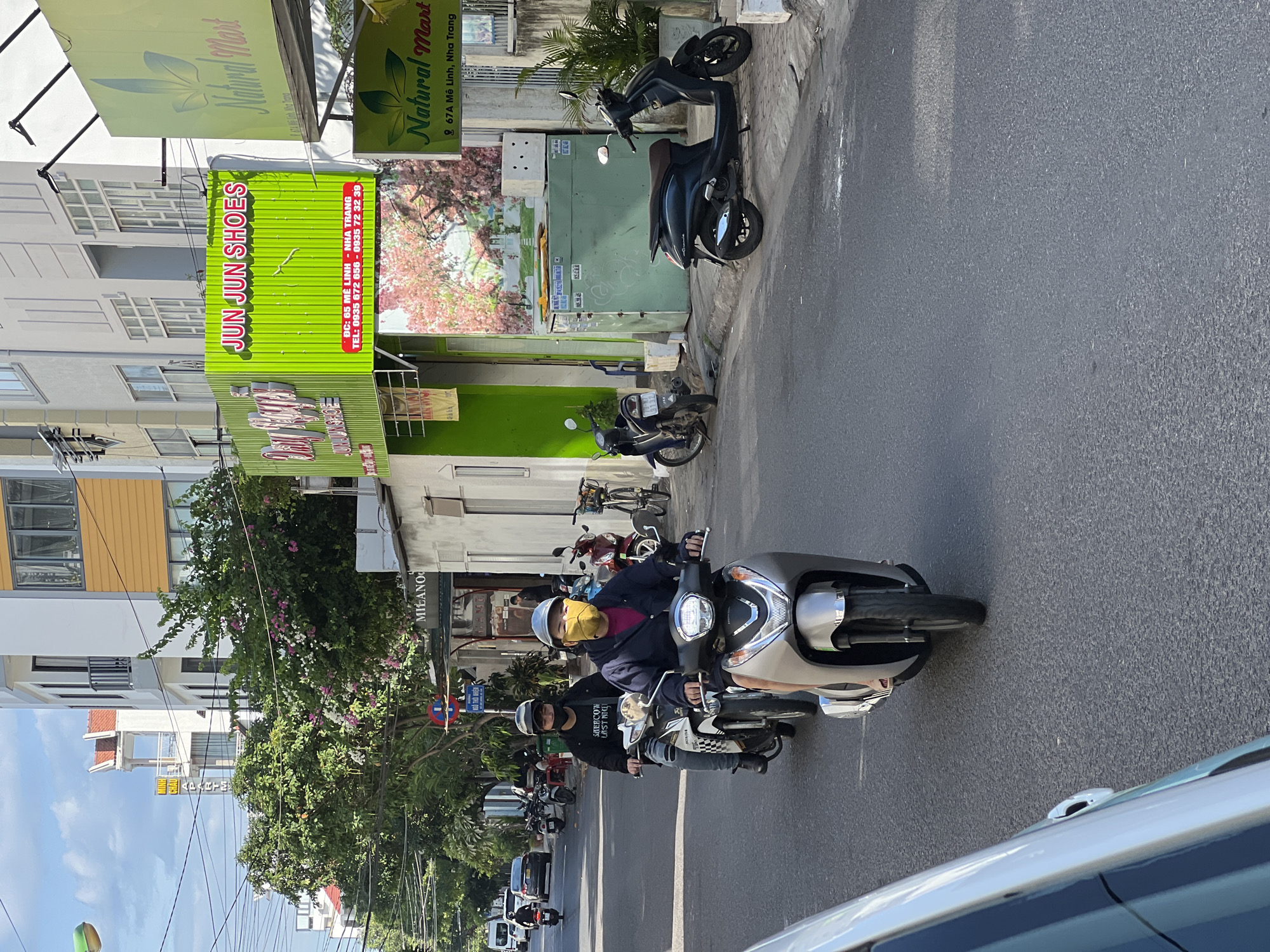 This is a street scene in Nha Trang, Vietnam. Two people are riding on a scooter in the foreground, with the driver wearing a gray helmet and a yellow face mask, and the passenger wearing a black helmet and a black face mask. They are riding on a paved road with buildings on the right side of the street. The buildings have signs for "JUN JUN SHOES" and "Natural Mart." There are other scooters parked on the sidewalk and a car in the distance. The sky is partly cloudy.