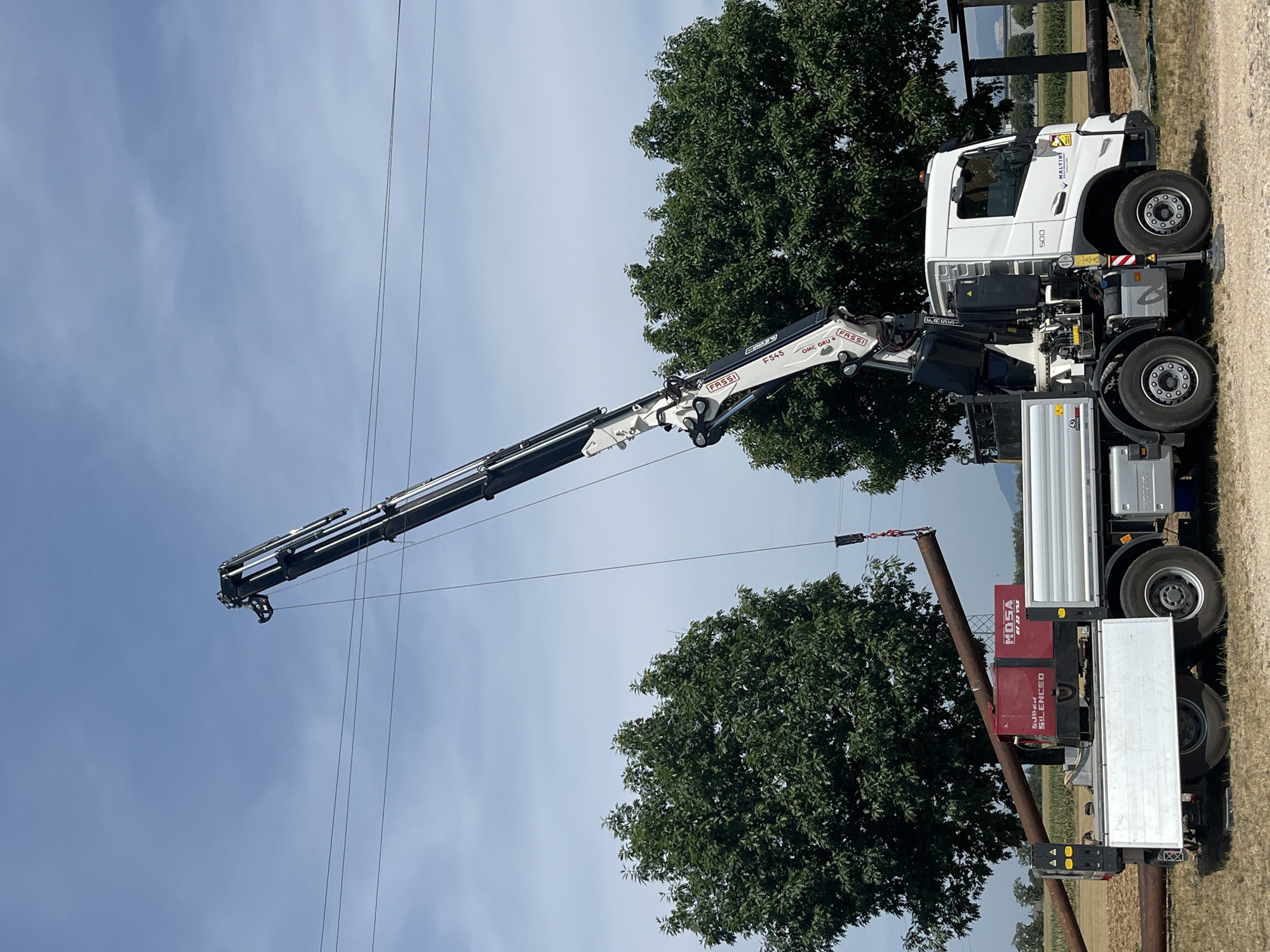 In Castiglione delle Stiviere, Italy, a white truck equipped with a crane is actively engaged in lifting a long, rusty metal pipe. The pipe is suspended from the crane's hook, positioned diagonally across the frame, and appears to be in the process of being loaded onto the truck's flatbed. Beside the truck's rear, a red generator with "MOSA SUPER SILENCED" branding is visible. The truck itself has a silver flatbed and various mechanical components associated with the crane. The scene is set outdoors under a partly cloudy blue sky, with lush green trees framing the background. Power lines are also present, stretching horizontally across the sky. The ground is a mix of dirt and gravel. No people are visible in the image.