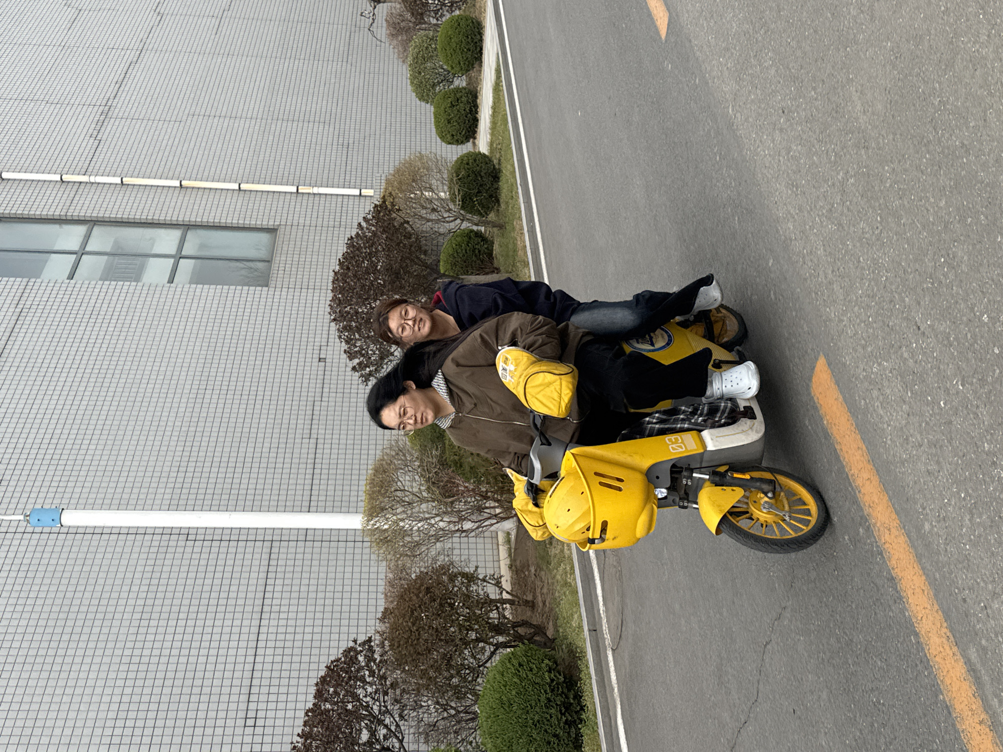 This image captures two individuals on a bright yellow electric scooter, likely a shared service vehicle, on an asphalt road in Zhongtun, China. The scooter is facing left from the viewer's perspective, with the road curving slightly to the left in the background.
The person in front, presumably the driver, is wearing a brown jacket over a checkered shirt, dark pants, and distinctive yellow gloves. They have dark hair and are wearing glasses, looking forward with a neutral expression. The second person is riding pillion, leaning slightly on the driver. They are wearing a dark blue jacket, jeans, and white Crocs, also wearing glasses and looking towards the camera with a slight smile.
The setting appears to be an urban or industrial area, characterized by a large, light grey building with a grid-patterned exterior on the left. A long, white horizontal light fixture is mounted on the building's facade. In front of the building and lining the side of the road are various bushes and shrubs, some bare-branched and some rounded and green, suggesting a temperate climate and possibly early spring. The road itself is asphalt with visible yellow and white painted lines.
Notable details include the scooter's vibrant yellow color and a number "60" visible on its side. The brand name "Meituan" (美团) is also visible on the front basket of the scooter. The image appears to be taken during the daytime under an overcast sky, as there are no harsh shadows. The overall impression is a casual moment of transit or rest on a shared scooter.