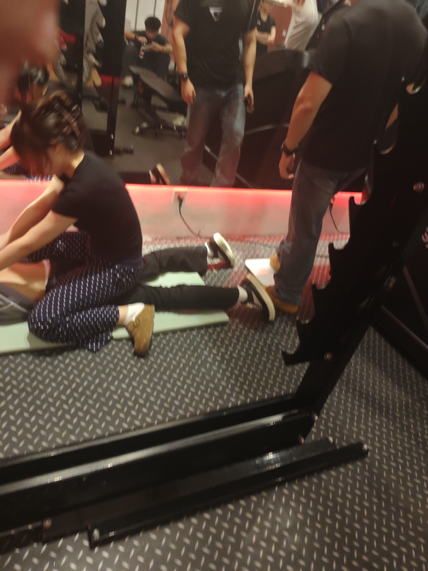 The image depicts an indoor gym environment located in the Chongqing High-tech Zone, China. In the central foreground, a woman with dark hair, pulled back, is kneeling on a light green exercise mat. She wears a black short-sleeved t-shirt, dark blue pants with a white dotted pattern, white socks, and brown slip-on clogs. Her hands are placed on the lower back/hip area of another individual who is lying prone on the mat, primarily visible by their legs in dark pants. This suggests a physical adjustment, stretch, or massage is being performed. To the right, a man stands with his back partially to the camera, wearing a black t-shirt, blue jeans, and brown shoes. The floor is composed of a grey metal diamond plate pattern. The background features a large mirrored wall reflecting the gym interior, including another individual in a black t-shirt and jeans standing by a counter or bench, and additional gym equipment. A horizontal red LED strip light is visible at the base of the mirrored wall, with an electrical outlet and a black cable plugged in. A black metal structure, consistent with gym equipment like a weight rack, is present in the lower right quadrant of the image.
