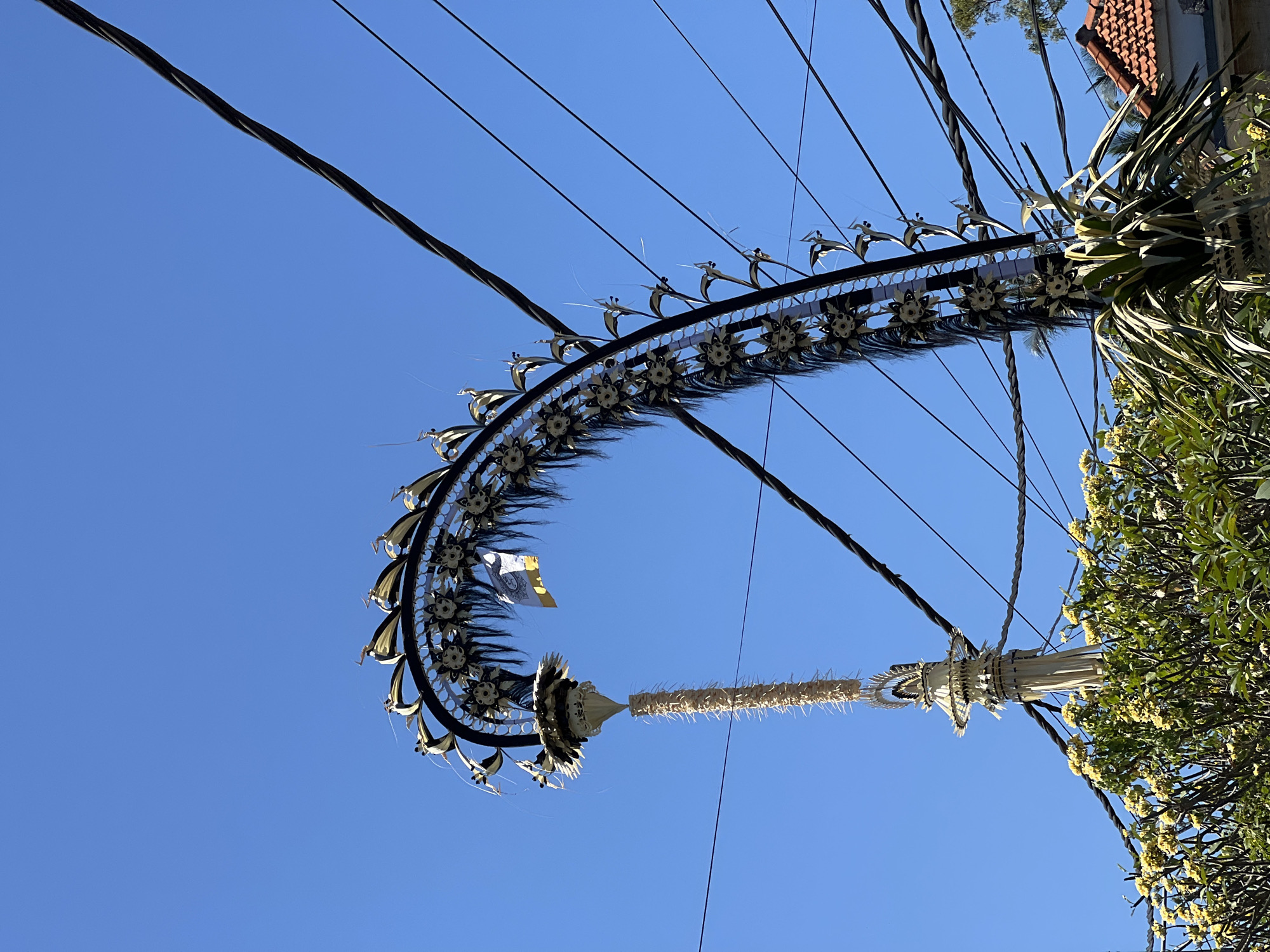 The photograph captures an outdoor scene in Manggis, Indonesia, dominated by a clear blue sky. In the foreground, a decorative structure, possibly a Balinese Penjor, is prominently displayed, curving from the left towards the right. This ornate structure is adorned with black tassels and decorative elements resembling flowers with gold accents. A long, woven straw-like hanging descends from the main structure, featuring spiky protrusions. The Penjor is positioned near a cluster of lush green foliage and blooming yellow flowers, suggesting a tropical setting. Several black electrical wires crisscross the upper portion of the image, creating a stark contrast against the bright sky. A small flag, yellow and white, is attached to the Penjor. In the background, the terracotta roof of a building is partially visible on the right side, hinting at residential or commercial structures in the vicinity. The scene is captured from a low-angle perspective, looking upwards. No people are visible in the image.