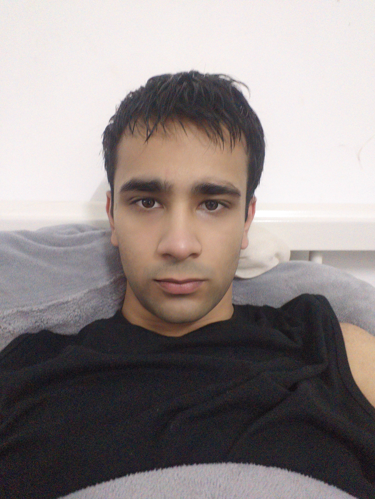 A man with dark hair and brown eyes is centered in a medium shot. He is wearing a dark, possibly black, t-shirt. He is resting against a light gray pillow or cushion, in front of a white wall. The image is taken in an indoor setting. There is no information to suggest the image was taken in Chongqing High-tech Zone, China.