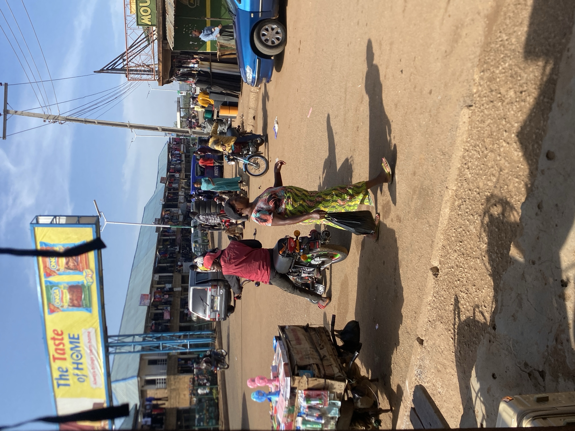 The image captures a vibrant, sunny street scene in Bukuru, Nigeria, likely a bustling market or commercial area. The photograph is taken from a slightly elevated perspective, looking down a thoroughfare populated with people, vehicles, and vendor stalls under a clear blue sky.
In the foreground, a woman in a colorful, patterned top and a green and yellow patterned skirt walks on the dirt road, casting a long shadow to her right. Adjacent to her, a person wearing a red shirt and a red cap is seated on a motorcycle, which has additional items strapped to its rear. In front of them, a wooden cart, likely belonging to a mobile vendor, is laden with various small goods, including brightly colored pink and blue plastic items.
The midground reveals more activity, with several individuals moving about, additional motorcycles, and a blue car parked by the roadside, displaying "H1972" on its side. A white van is also visible further down the street. Buildings housing various stalls and shops line both sides of the street, indicating a lively commercial environment.
In the background, a tall utility pole with numerous overhead wires stretches into the sky. A large yellow billboard on the left prominently displays the text "The Taste of HOME" and shows packages, one clearly marked "ONGA." Another partial sign at the top right reads "MOU...". The strong, distinct shadows cast by people and objects suggest bright sunshine, indicating it is likely mid-morning or afternoon. The overall atmosphere is busy and active, reflecting daily life in a lively urban or semi-urban setting.