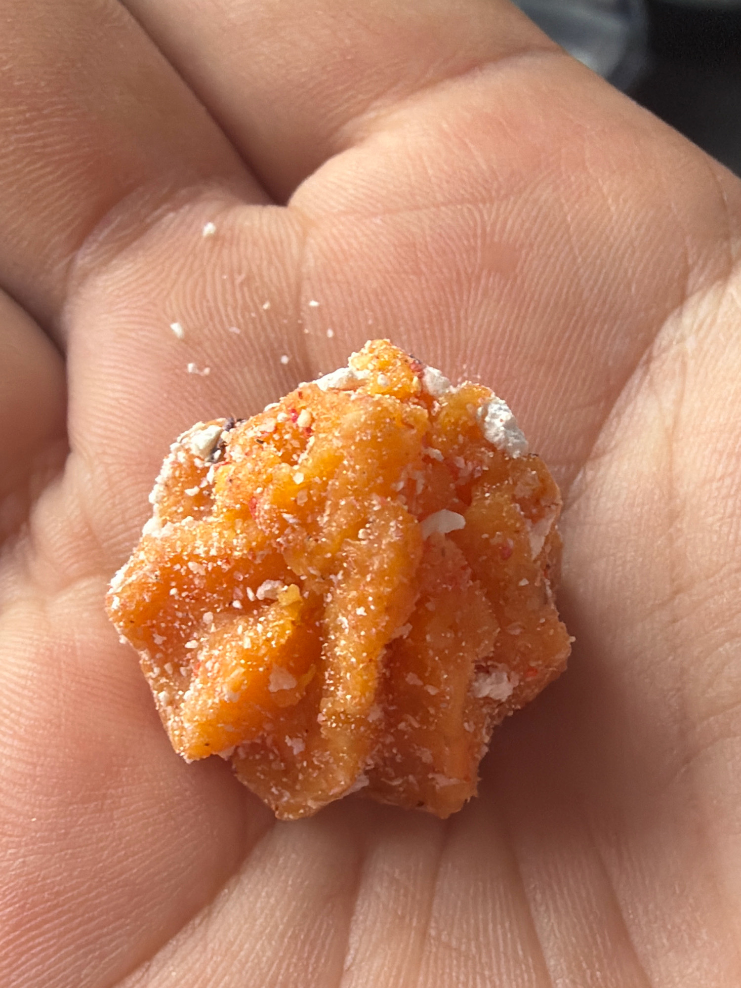 The image is a close-up shot of a person's hand holding a piece of orange food, possibly candy or a dried fruit, covered in white sugar crystals. The food item is irregular in shape and appears to be made of compressed pieces. The hand is lightly textured with visible fingerprints and skin creases. The background is out of focus, showing hints of a lighter tone on the right. The location in Бактуу-Долоноту, Kyrgyzstan is not discernible from the image itself.