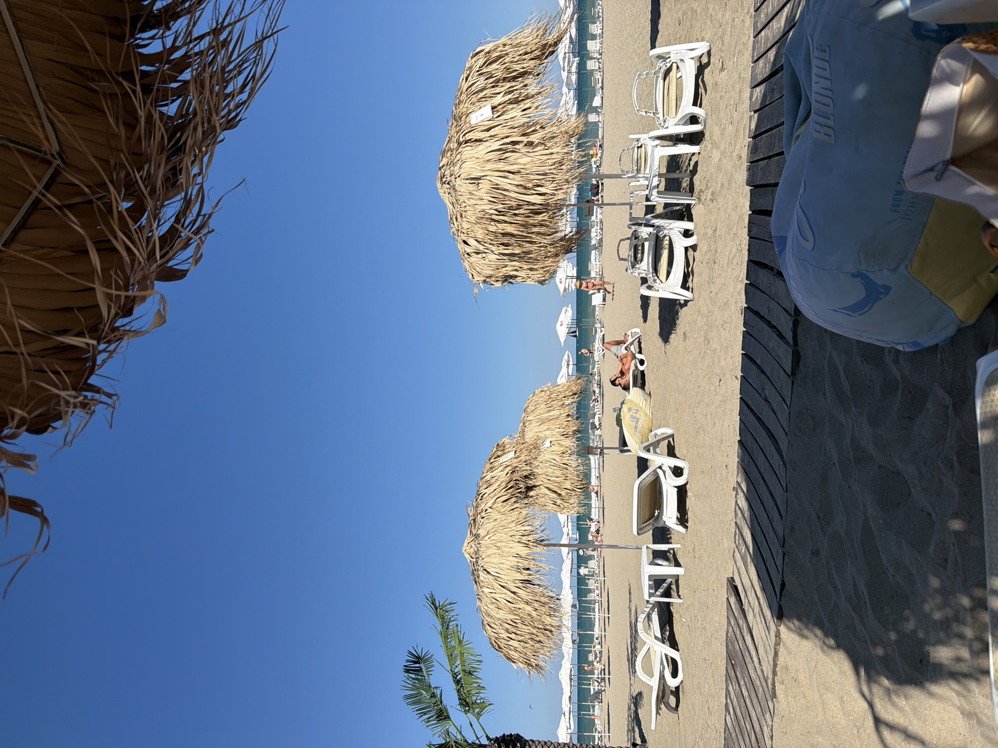 The image depicts a sunny day at a beach, likely in Burgas, Bulgaria, characterized by its sandy shores and the clear blue sky. In the foreground, a large, light blue beanbag chair with the word "BLONDE" printed on it rests on the sand, partially obscuring a wooden boardwalk. Beyond the boardwalk, the beach is dotted with numerous white lounge chairs and straw-thatched umbrellas, some occupied by sunbathers. A palm tree is visible on the left side of the frame. The sea stretches out in the background, with a few distant beachgoers visible. The scene conveys a relaxed atmosphere of a typical summer day at the seaside.