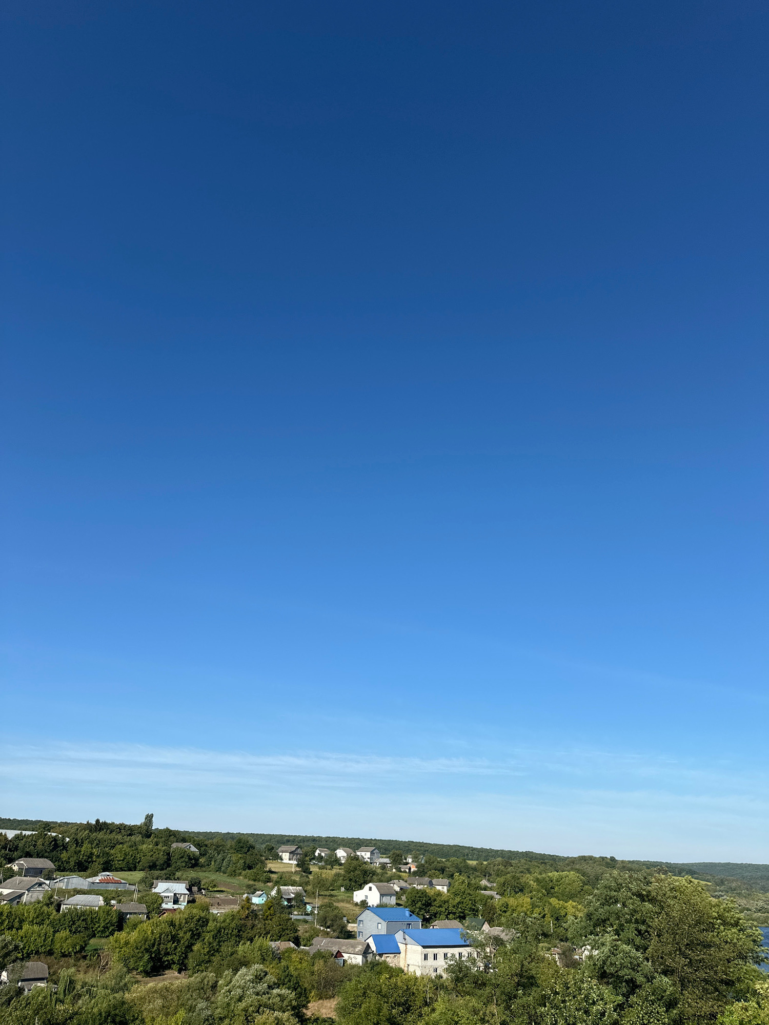 The image depicts a clear, bright blue sky dominating the upper portion, with a vast expanse of uninterrupted sky. Below the sky, a rural landscape unfolds. A cluster of houses with predominantly grey and blue roofs are scattered amongst lush green trees and rolling hills. The architecture suggests a residential area, possibly a village or a small town. In the far distance, a body of water, likely a river or lake, is visible on the right side, reflecting the blue of the sky. The overall scene conveys a peaceful, daytime setting in a natural environment. The image location is Makiv, Ukraine.