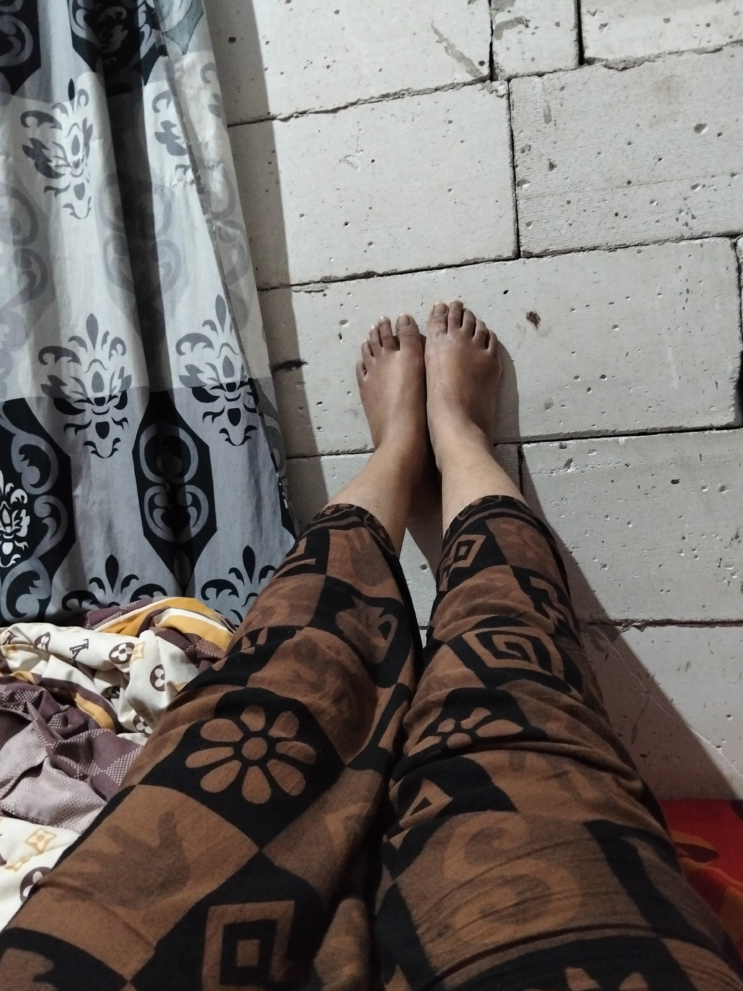 The image shows a pair of bare feet resting on a pile of blankets, with the legs clad in brown patterned pants. In the background, a white brick wall is visible, with a portion of a gray and black patterned curtain on the left side. The scene appears to be indoors, possibly a bedroom, given the presence of blankets and curtains. The lighting suggests it is either evening or a dimly lit room. There are no other people or significant objects in the frame besides the feet, legs, pants, blankets, wall, and curtain. The location in Panimbang, Indonesia, is not evident from the image itself.