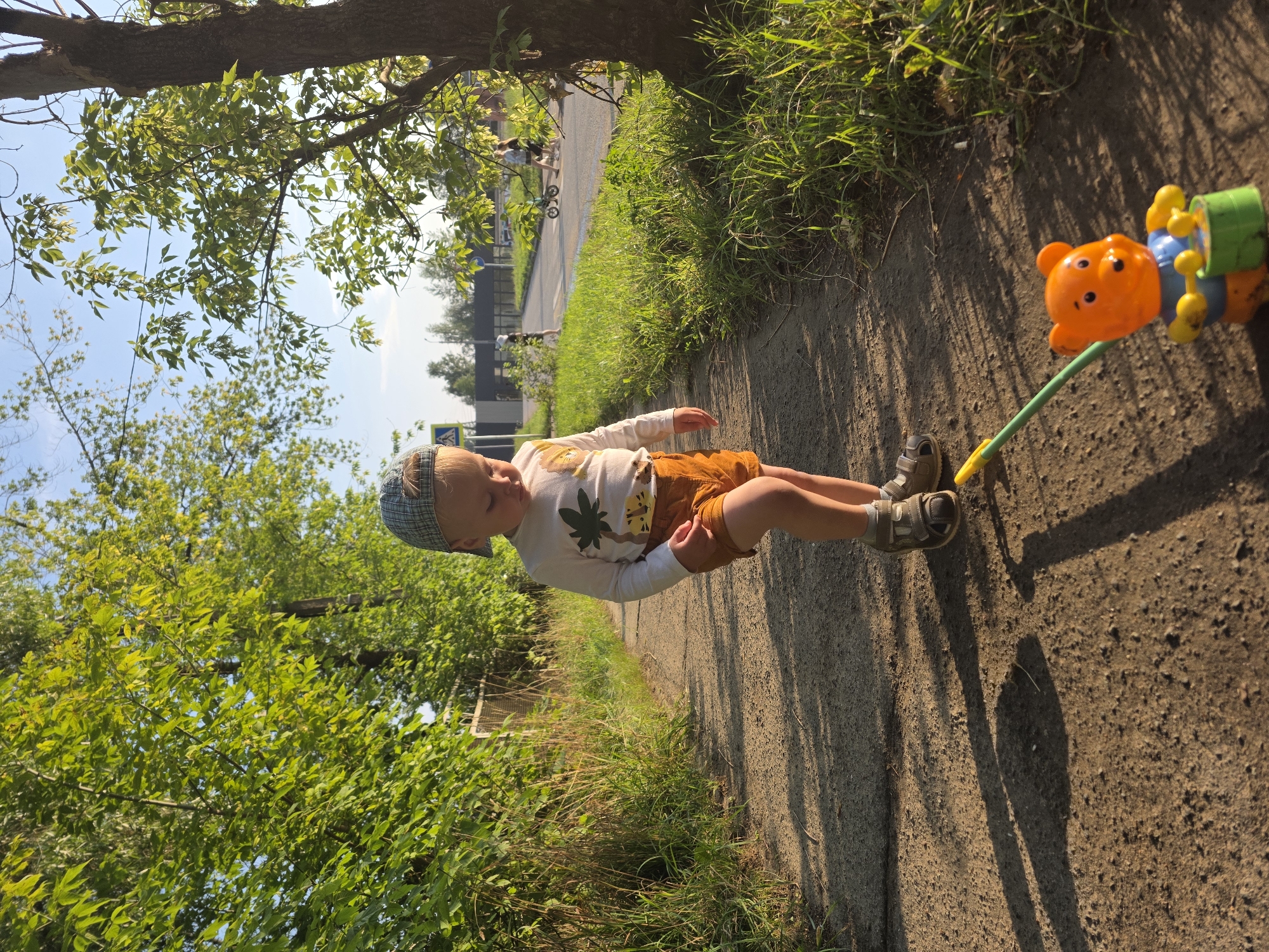 A young child wearing a plaid cap, a long-sleeved white shirt with a palm tree and tiger print, and orange shorts walks on a paved path. The child is looking down at a toy bear on wheels with a green bucket. The path is bordered by green grass and trees. In the background, there is a road with several people and a bicycle. The scene appears to be a residential street in Chaikovsky, Russia, on a sunny day.