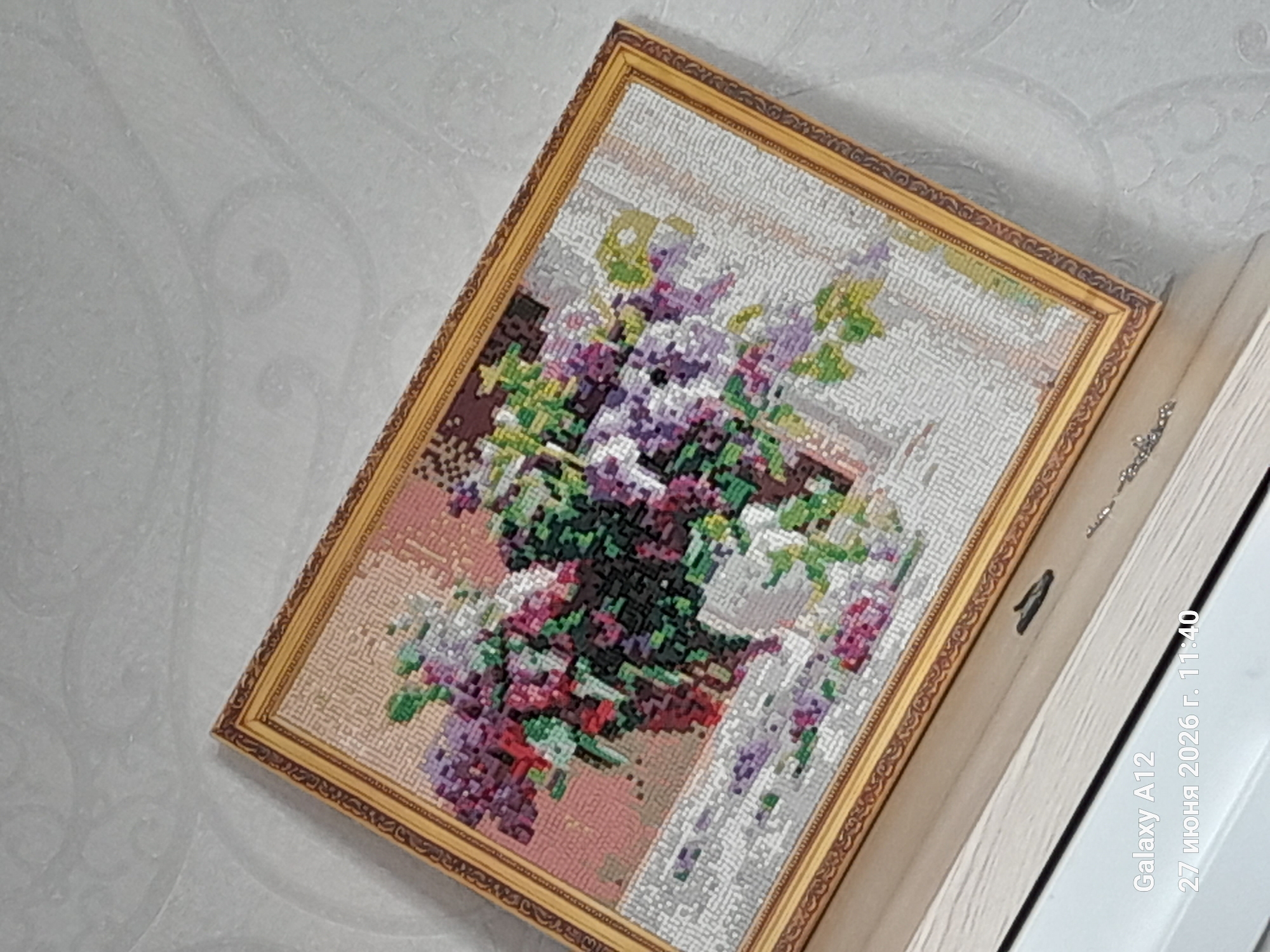 This image shows a framed needlepoint artwork depicting a vase of lilacs on a table, placed against a textured wallpaper background. The artwork is a detailed representation of lilacs in various shades of purple, pink, and green, with a brown vase and a white tablecloth. The frame is ornate and golden. The image also captures a portion of a shelf or surface the artwork is resting on, which appears to be made of wood. In the bottom left corner, a text overlay from a device indicates "Galaxy A12" and "27 июня 2026 г. 11:40" which translates to "June 27, 2026, 11:40". This information, especially the date, is likely metadata from the device used to take the picture and does not relate to the actual content of the artwork or its environment. The specific location in Kara-Köl City, Kyrgyzstan is not visually indicated within the image itself, only through the prompt's context. The lighting suggests an indoor setting, possibly a home.
