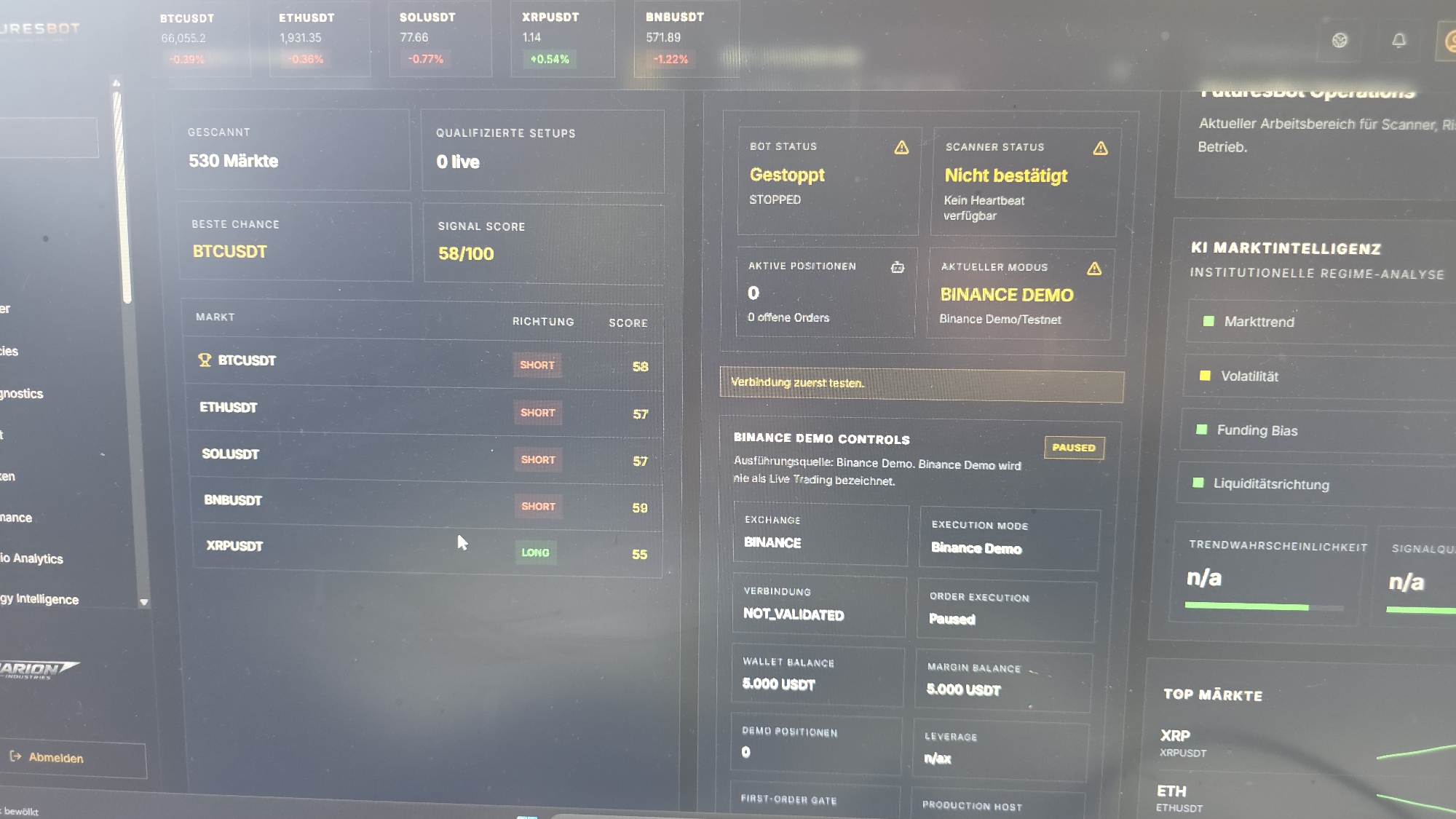 The image shows a screenshot of a cryptocurrency trading platform called FuturesBot. The platform displays various market information, bot status, and trading controls. On the left side, there's a list of markets like BTCUSDT, ETHUSDT, SOLUSDT, BNBUSDT, and XRPUSDT, with their respective scores and trading directions (SHORT or LONG). The central panel shows the bot status as "Stopped" and the scanner status as "Not confirmed" with a warning icon. It also indicates that the current mode is "Binance Demo" and there are 0 active positions. The right panel details "KI MARKTINTELLIGENZ" (AI Market Intelligence) with indicators for "Markttrend" (Market Trend), "Volatilität" (Volatility), "Funding Bias," and "Liquiditätsrichtung" (Liquidity Direction), all showing as green or yellow squares, suggesting positive or neutral states. The platform also shows connection status and wallet balance for the Binance Demo account. There is no information in the image that relates to Hirtenberg, Austria.