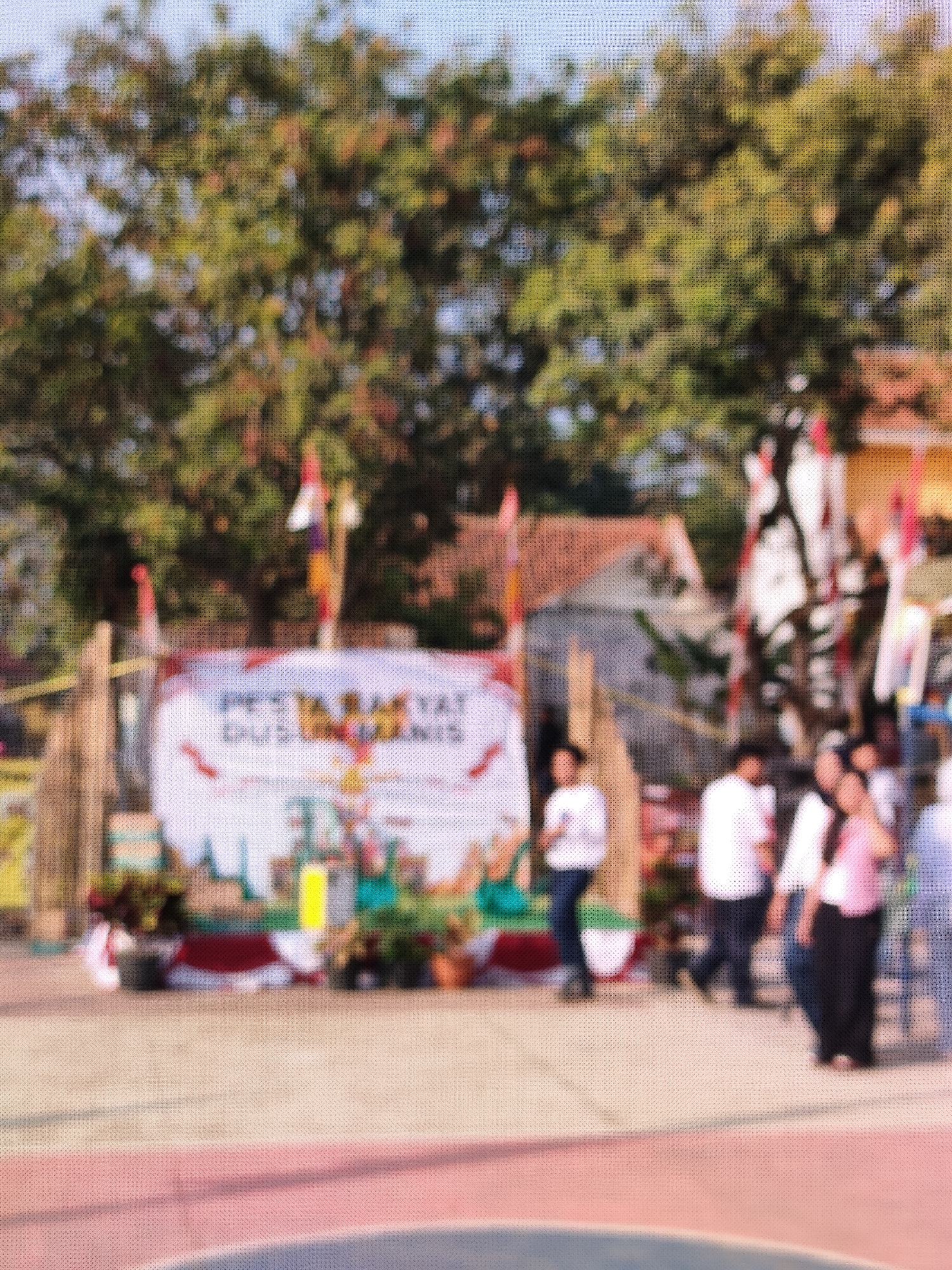 The image depicts an outdoor scene with a banner in the background, likely at an event. The banner is white with red and blue lettering and graphics, including an image resembling the Garuda Pancasila, the national emblem of Indonesia. Several people are visible in front of the banner, some walking and others standing. One individual, wearing a white shirt and dark pants, stands prominently in the center. To the right, a person in a pink shirt and dark pants is walking. The environment includes trees and buildings in the background, suggesting a public space or park. The foreground shows a paved surface with colored lines, possibly a sports court or walkway. The image is heavily blurred, making precise identification of details difficult. The prompt's request to specify the location in "the city of Caracas, Indonesia" is contradictory, as Caracas is the capital of Venezuela and not located in Indonesia. Therefore, the location within Indonesia cannot be determined from the provided image.
