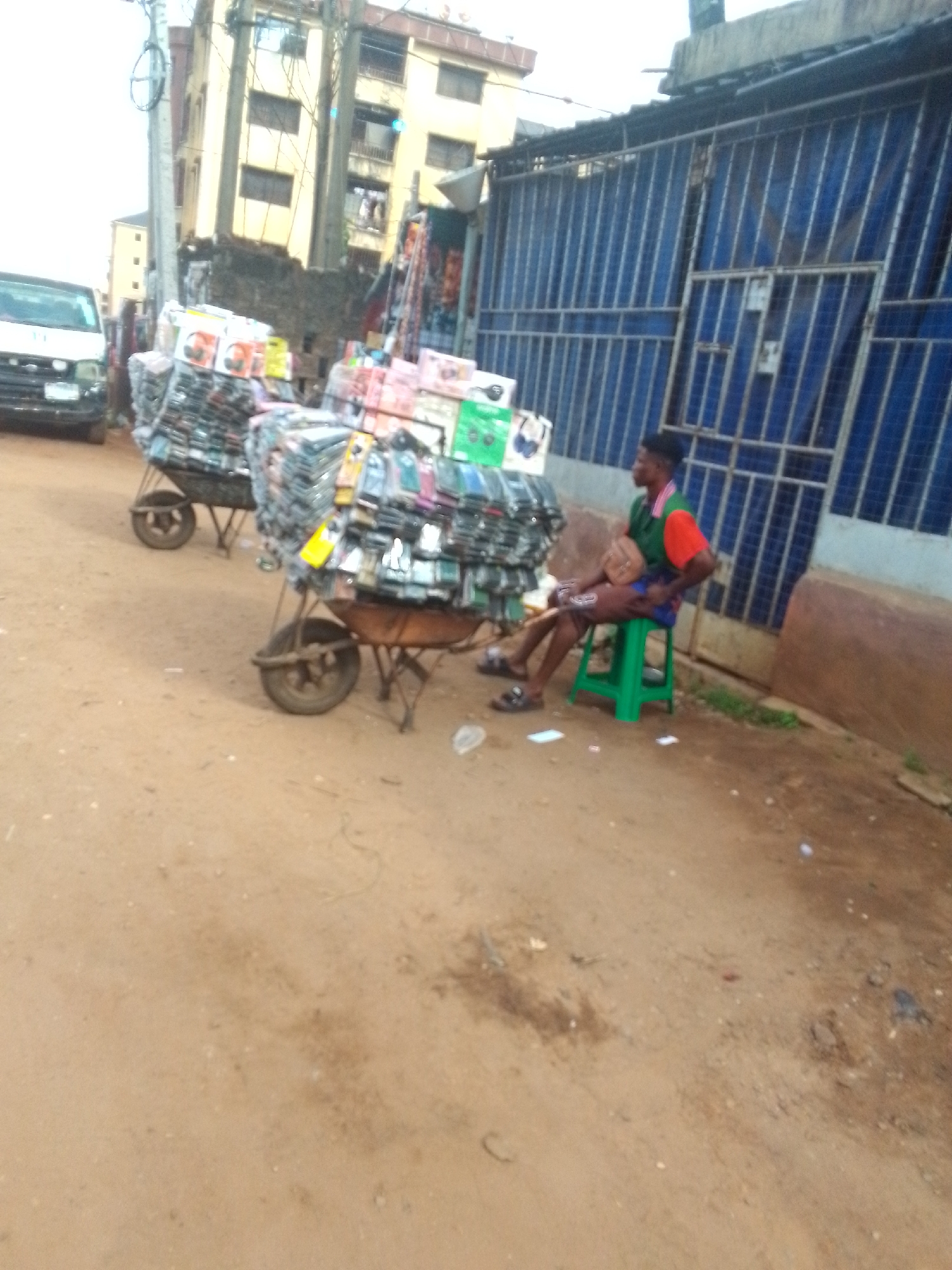 In Onitsha, Nigeria, a man sits on a green stool near a building with blue metal gates. Two wheelbarrows are laden with goods, including mobile phone cases and headphones. A white van is parked nearby. The ground is dirt.