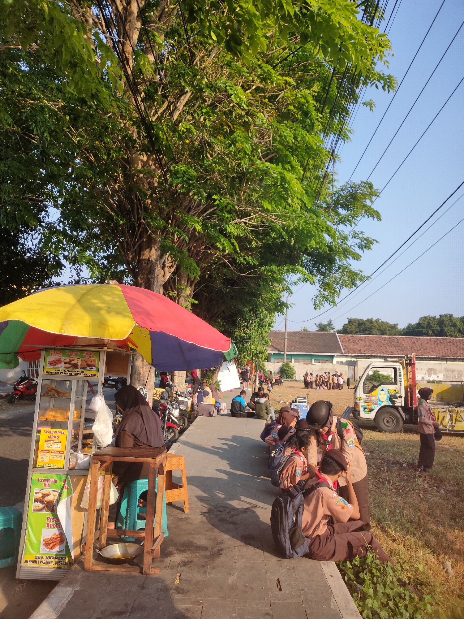 In the city of Ngasem, Indonesia, a street-side vendor with a colorful umbrella sells snacks next to a pathway. Several people, some wearing scout uniforms, are gathered in small groups along the pathway and on the grass. A white truck is parked nearby, and a group of children or young people stand in front of a building in the background. Trees line the street, with power lines running overhead against a clear blue sky. The scene appears to be a casual gathering or event in an outdoor public space.