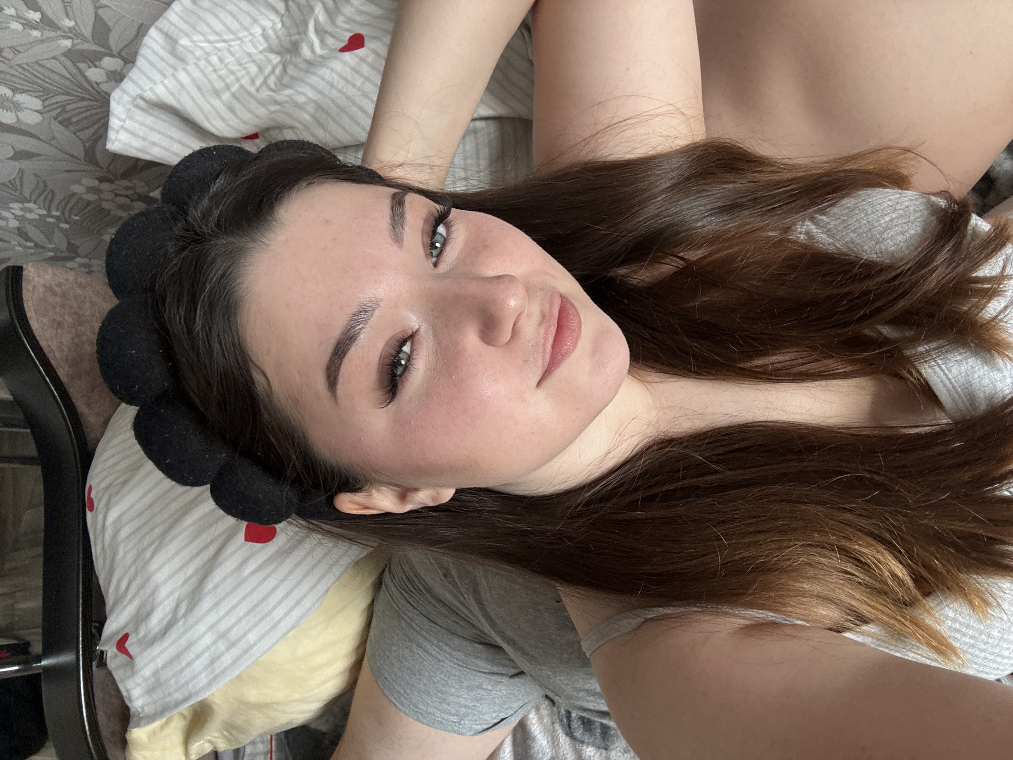 The media file shows a close-up, overhead shot of a young woman lying on a bed. She is wearing a grey t-shirt and a black headband with soft, rounded protrusions. Her long, dark brown hair is spread across the bed and her shoulders. She has light skin, green eyes, and is looking directly at the camera with a neutral expression, though her slightly parted lips suggest a subtle smile. Her makeup includes defined eyebrows and long eyelashes.
The background consists of a striped pillow with small red heart accents, and what appears to be a floral patterned bedsheet. A yellow pillow is partially visible beneath the striped one. The lighting suggests daytime, possibly morning or afternoon, with soft, diffused light. The overall impression is a casual, relaxed moment, likely captured at home. No visible text is present in the image.