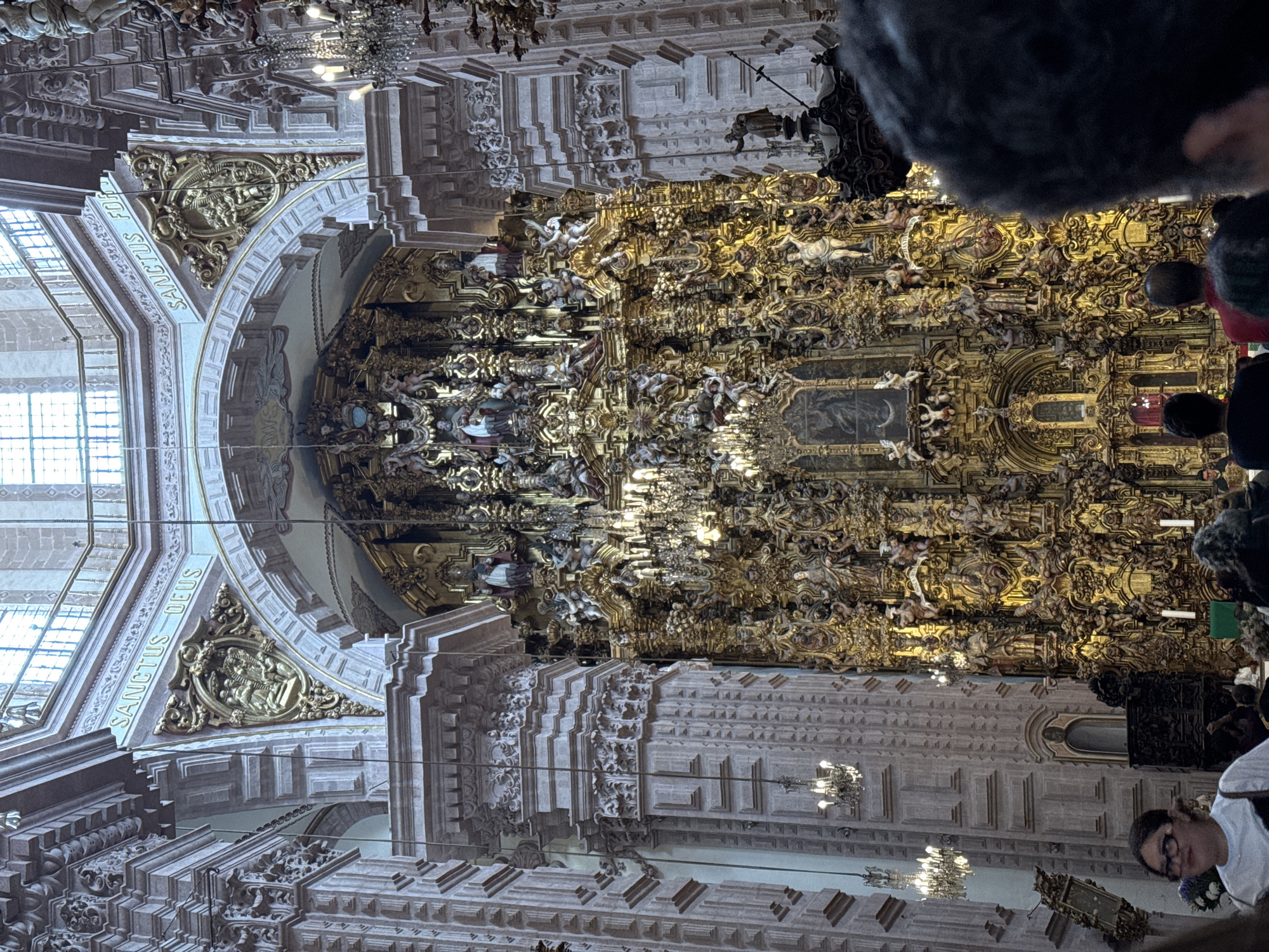 The image depicts the interior of the Santa Prisca Church, located in Taxco de Alarcón, Guerrero, Mexico. The central focus is a massive, multi-tiered Churrigueresque altarpiece (retablo) densely covered in gold leaf, occupying the front of the nave. This elaborate structure features numerous sculpted figures of saints, angels, and cherubs, intricate carvings, decorative columns, and cornices. A painting is visible within a gilded frame at the lower-center of the retablo, flanked by candlesticks.
Above the altarpiece, within the architectural framework, large arched openings reveal upper windows providing natural light. Inscribed Latin phrases, "SANCTUS DEUS" (Holy God) and "SANCTUS FORTIS" (Holy Mighty), are visible on the arches to the left and right, respectively.
The church's interior architecture is characterized by monumental columns and pilasters with deeply carved, textured surfaces and ornate capitals. Multiple ornate chandeliers with lit bulbs hang from the ceiling and project from the side walls, illuminating the space. The upper walls and ceiling exhibit painted decorations and intricate relief work.
In the foreground and mid-ground, the backs and tops of several individuals' heads are visible, facing towards the main altar, suggesting a congregation or visitors observing the scene. In the lower-left, a woman with glasses and dark hair, wearing a white shirt, is partially visible, smiling slightly and looking towards the left. In the lower-right, the back of a man's head with dark, curly hair is prominent, partially obscuring the view of the altar.