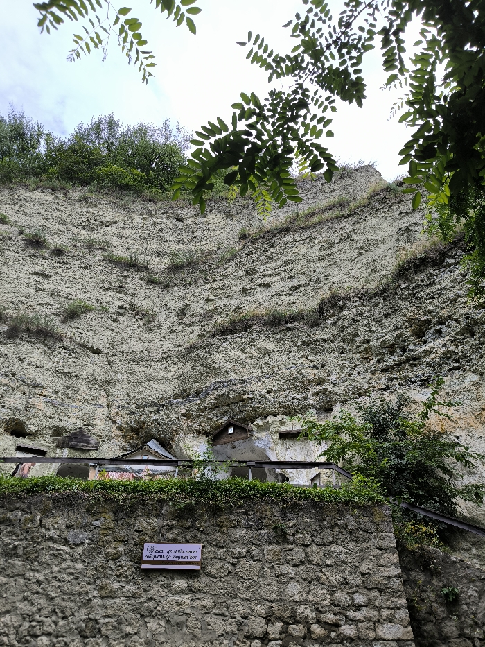 This image depicts a detailed view of a monastic complex built into a natural cliff face in Horaivka, Ukraine. The scene is dominated by a towering, light-colored cliff composed of layered rock, exhibiting erosion and sparse green vegetation, including grasses and small shrubs, growing directly on its surface.
At the base of the cliff, several structures are visible. These include small, simple buildings with sloped roofs, seemingly integrated into natural caves or carved directly into the rock face. One notable structure has a brown-pitched roof with a dark, rectangular opening beneath it. To its right, another dark, rectangular opening is present in the rock. Horizontal wooden or metal beams or railings extend along the front of these structures, indicating a possible walkway or supporting elements.
In the foreground, a robust stone wall, constructed from irregularly shaped, light-colored stones, serves as a retaining structure or boundary. Above this wall, a line of dense green foliage runs horizontally, possibly on top of a railing or fence that is partially obscured. Affixed to the center of this stone wall is a white rectangular sign with dark Cyrillic text that reads: "Душа не може довго Собирать до многово Бог." (The soul cannot long gather to the many God.)
The top portion of the image shows a bright, overcast sky with some visible clouds. Overhanging branches with green leaves are present in the upper left and right corners, partially obscuring the sky and upper cliff edge. Denser green trees and bushes are visible along the top edge of the cliff. No individuals are present in the scene.