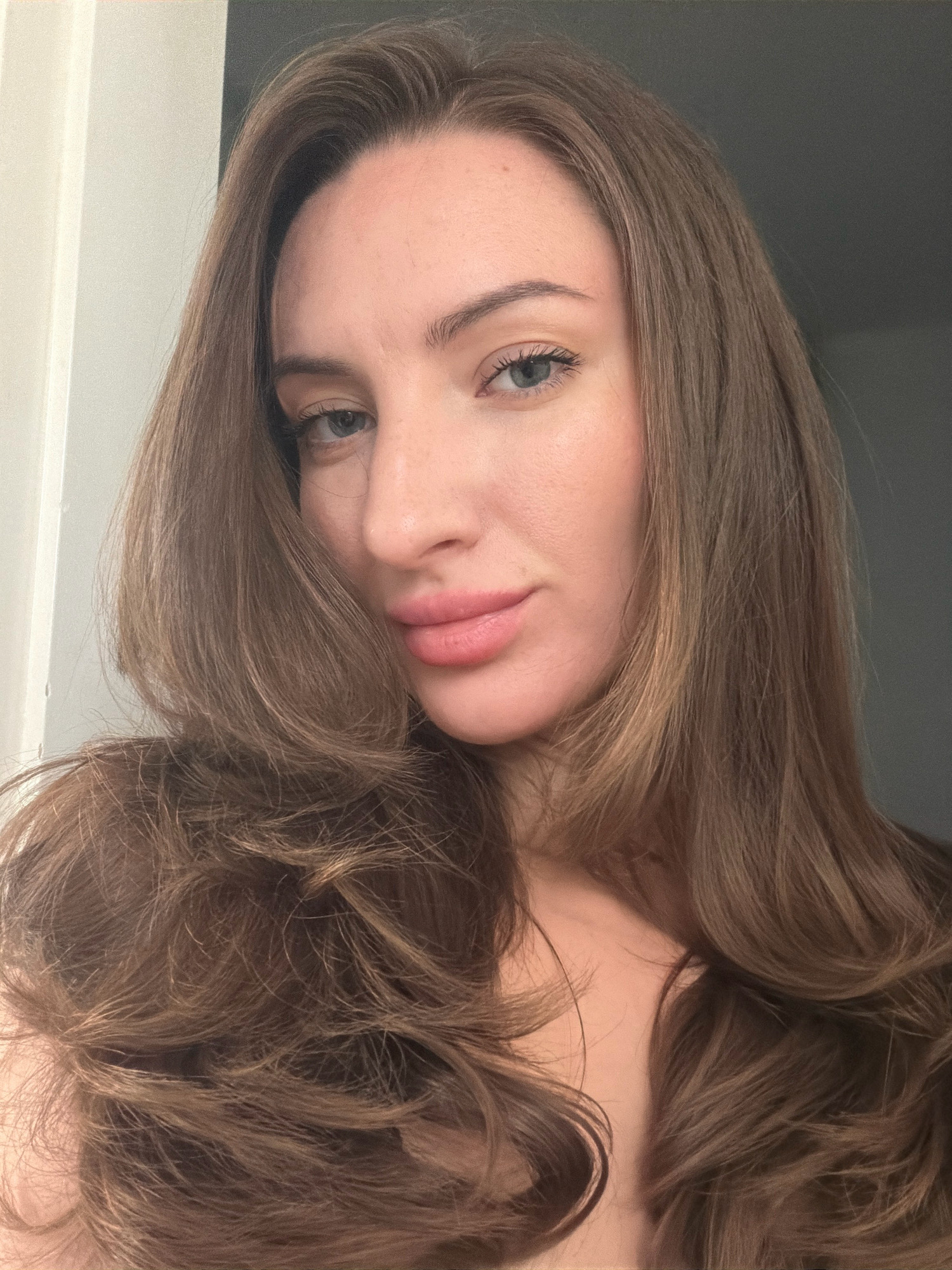 A close-up, head-and-shoulders selfie of a woman with long, brown hair framing her face. She has fair skin, light blue eyes, and pink lips. The background is indistinct, with a light-colored wall on the left and a darker, possibly grey, surface on the right. There is no indication of the location being Sharm El Sheikh, Egypt.