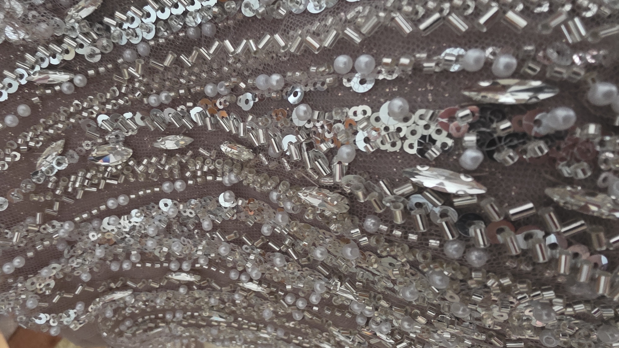 The image shows a close-up of a densely beaded and sequined fabric. The base fabric appears to be a pale, possibly nude or mauve-toned netting. The embellishments consist of various types of beads: small round pearls, cylindrical silver beads, and silver sequins. Larger, clear, faceted stones are also scattered throughout. The beads are arranged in irregular patterns, some forming lines. The environment is unknown as the image is a macro shot of a fabric surface. The location in Blagoevgrad, Bulgaria, is not determinable from the visual information.