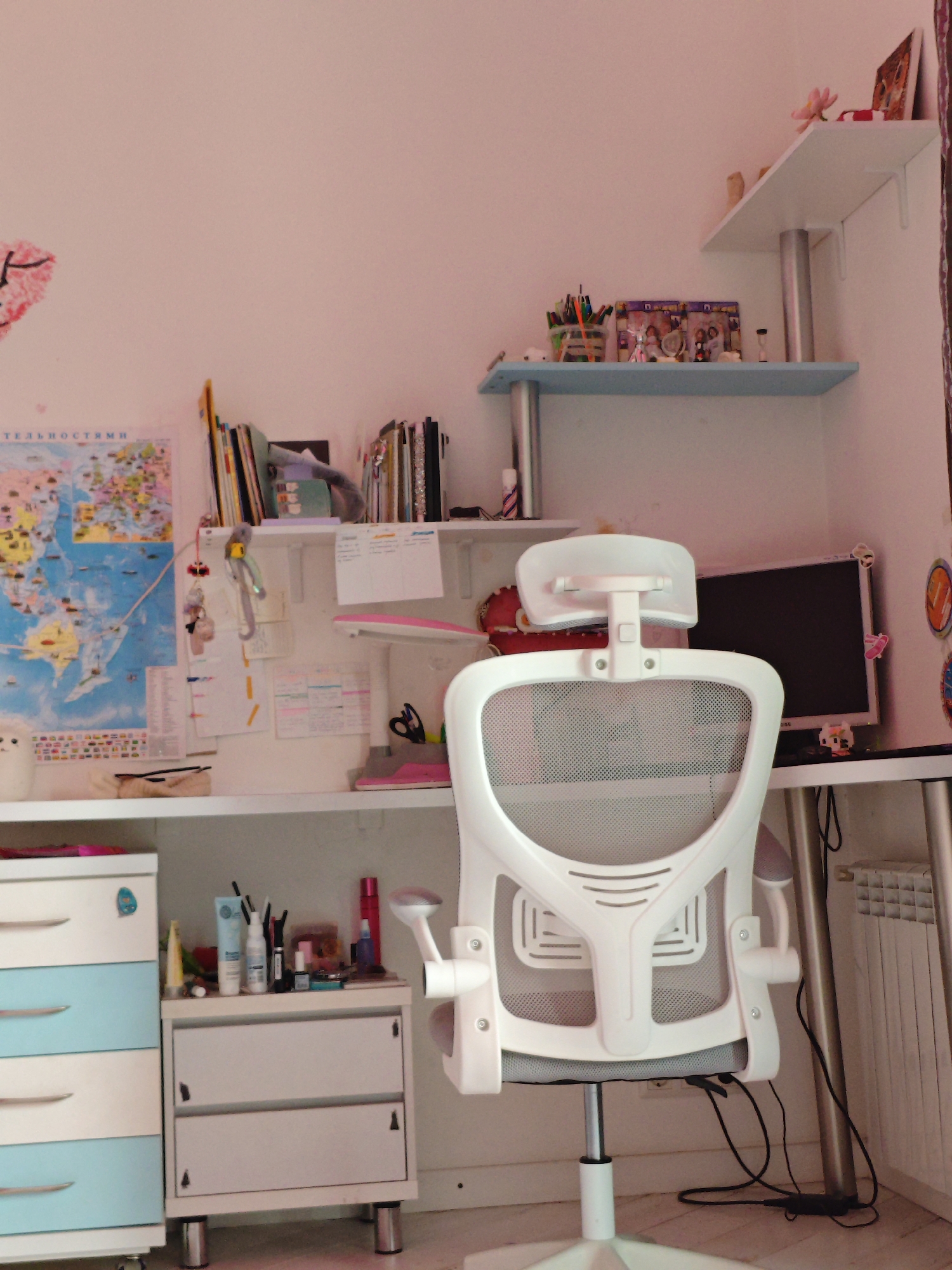 This is a forensic, matter-of-fact description of a desk area, likely in a residential setting, with no clear indicators placing it specifically within Трудовское сельское поселение, Ukraine.
The scene depicts a white ergonomic office chair with a mesh back and seat positioned in front of a desk. The desk is cluttered with various items, including a computer monitor, pens, pencils, and what appear to be art supplies. Shelves above the desk are also filled with books, decorative items, and more art supplies. A world map is pinned to the wall behind the desk, alongside various notes and papers. To the left of the desk is a chest of drawers with light blue and white fronts. To the right, a radiator is visible. The overall environment suggests a workspace or study area, with a slightly disorganized but functional arrangement of objects. There are no people present in the image.