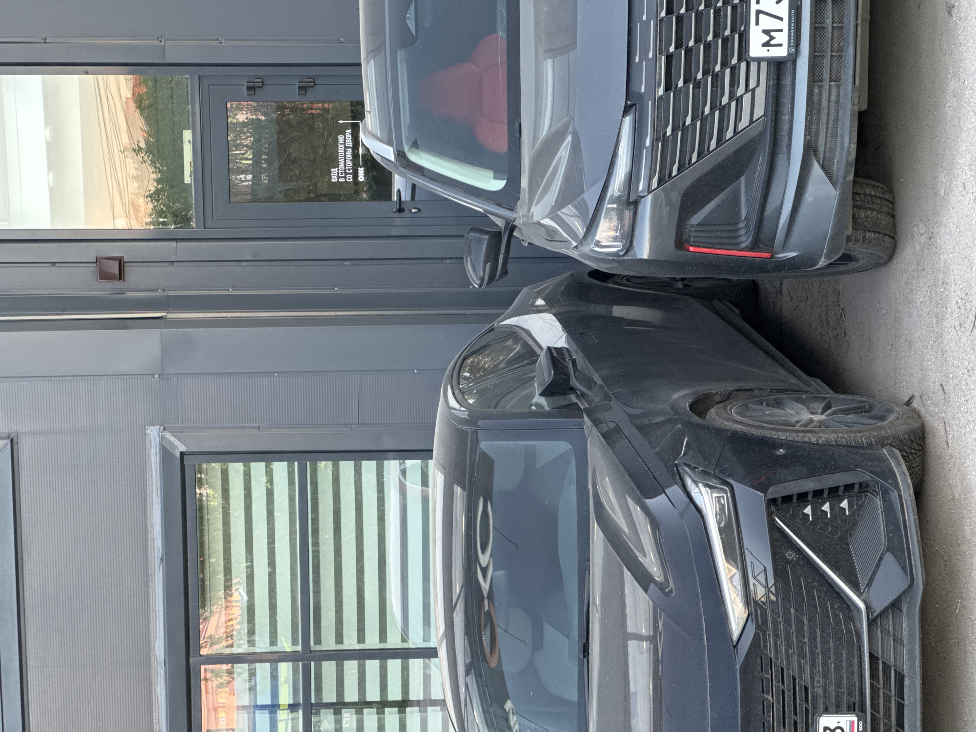 The image shows the front ends of two dark-colored cars parked close together. The car on the right is grey and has a license plate ending in "M73". The car on the left is black and has a license plate ending in "83". The scene is in front of a building with a glass door and a window. The door has a sign in Russian that includes the words "СТОМАТОЛОГИЮ" (stomatology) and "СО СТОРОНЫ ДВОРА" (from the yard). The location is in an urban area, possibly near a dental clinic. The city and country are unknown.