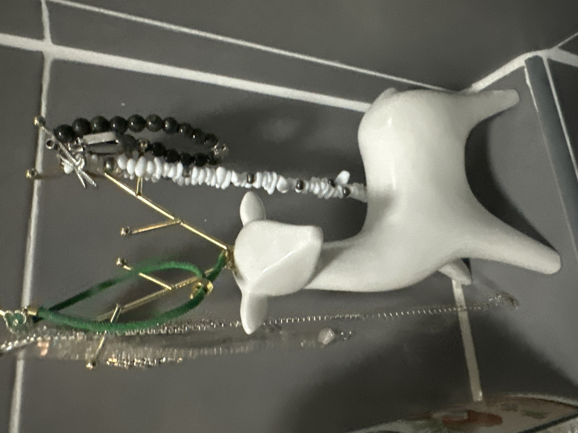 A white ceramic deer figurine holds several necklaces on its antlers. The figurine is positioned on a shelf against a grey tiled wall. To the left, a silver chain necklace with small clear beads hangs. A green cord necklace with a small green charm is also visible. To the right, two beaded bracelets hang, one with white beads and the other with dark grey beads. The background appears to be a bathroom or vanity area. The specific location in Kathu, Thailand is not ascertainable from the image.