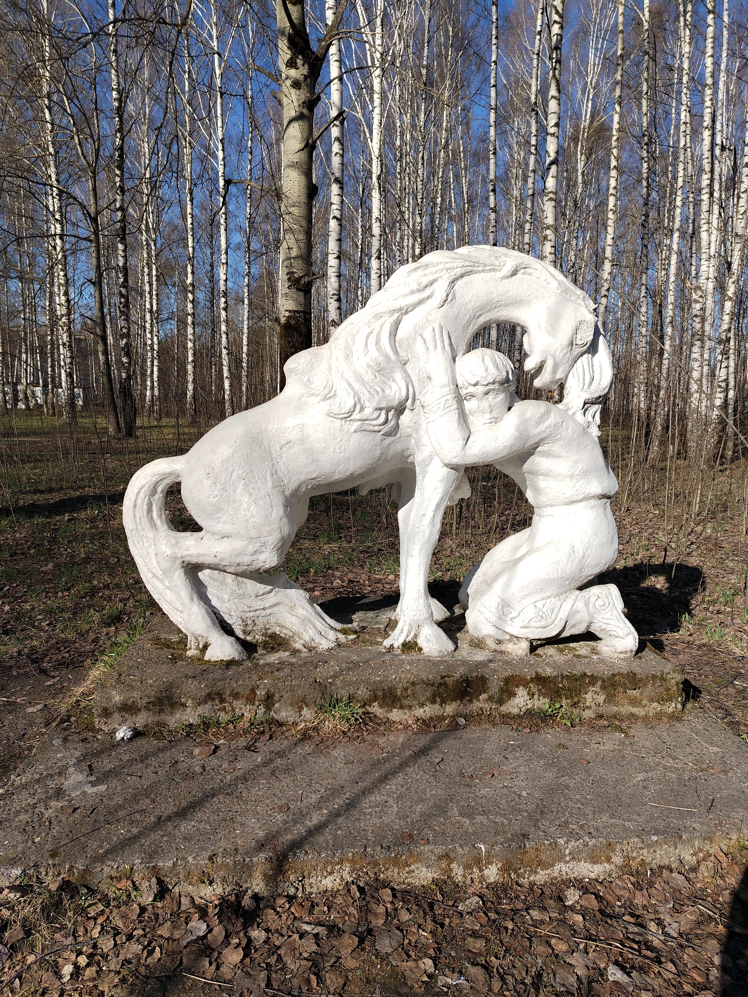The image depicts a white plaster sculpture of a kneeling person embracing a horse. The sculpture rests on a weathered concrete base with some moss growth. The background is a forest with numerous tall, slender birch trees with white bark. The sky is blue and visible through the branches. The location is likely in Chaikovsky, Russia, given the prompt's request. The ground around the base is covered in fallen leaves and sparse vegetation. The sculpture is the primary subject, showing a human-animal interaction. The setting is outdoors, likely in a park or wooded area.