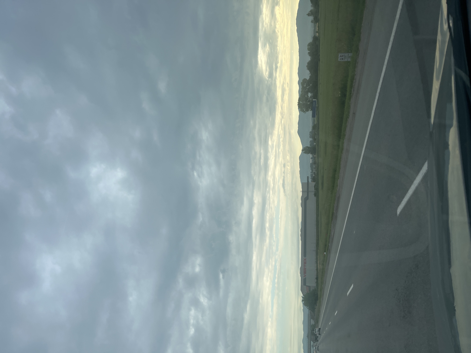 A long shot captures a highway stretching into the distance under a cloudy sky. On the right side of the road, a vast green field is bordered by trees and mountains in the background. A large building with "VERTEX" in red letters is visible on the left side of the road. The perspective suggests the photo was taken from a moving vehicle, as evidenced by the dashboard in the foreground and the dashed white lines on the asphalt. The lighting is overcast, suggesting it may be dawn or dusk. There are no people or animals visible in the frame.