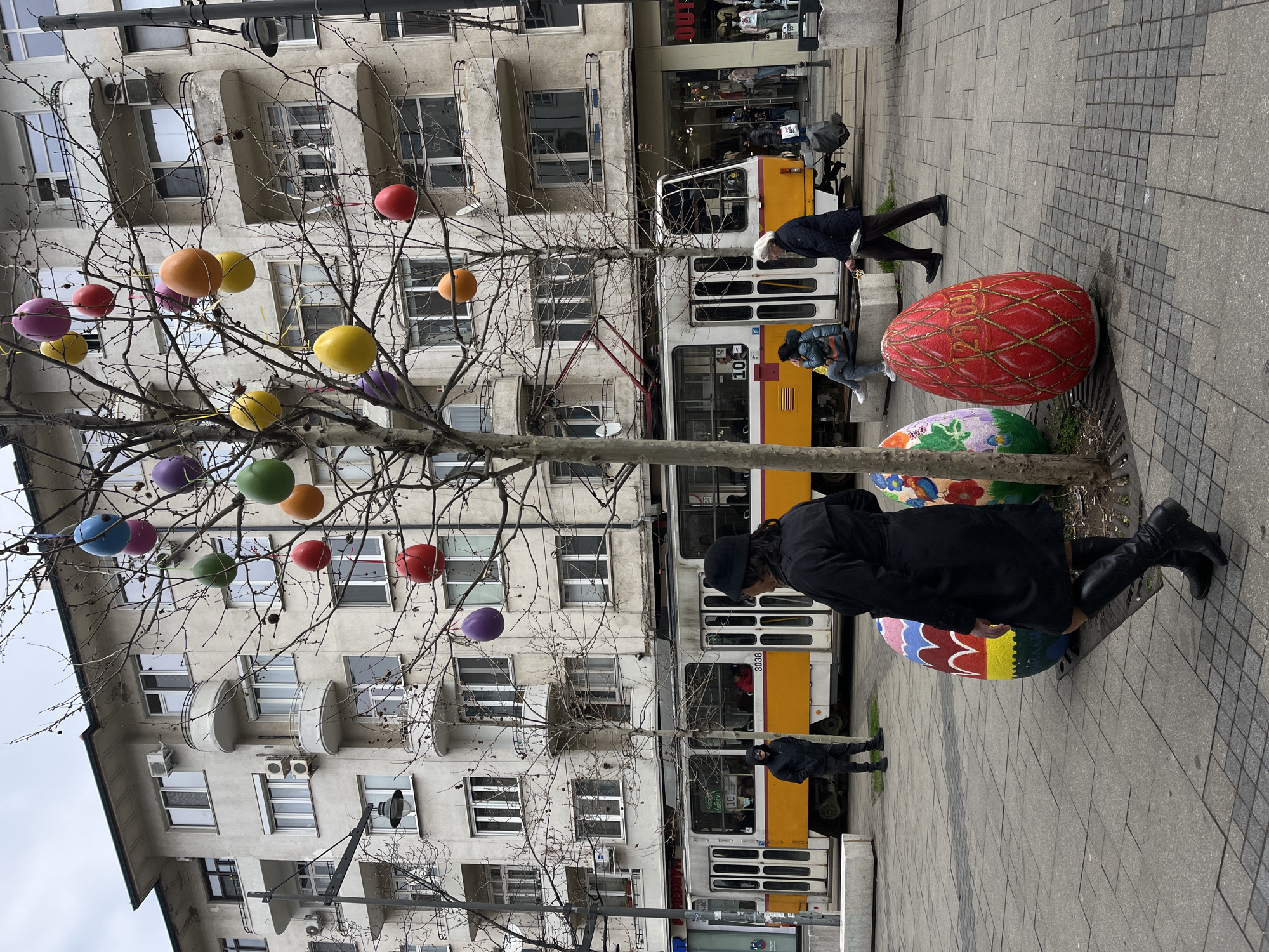 The image depicts a street scene in Sofia, Bulgaria. In the foreground, there is a tree decorated with colorful spherical ornaments, resembling Easter eggs. Adjacent to the tree base are several large, painted Easter egg sculptures on the sidewalk. A person dressed in a black coat and boots is bending down near the eggs. In the background, a white and yellow tram is visible, along with pedestrians walking along the sidewalk. A multi-story building with numerous windows is situated behind the tree and tram. The sky appears overcast.