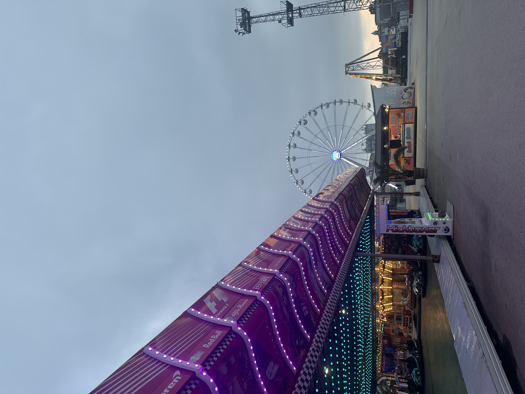 This image depicts an amusement park or fairground in Varna, Bulgaria, under an overcast sky. In the immediate foreground to the left, a bumper car ride structure is visible. Its curved roof canopy is predominantly reddish-purple, adorned with wavy lines of glowing purple LED lights. Along the top edge of this canopy, various Formula 1 brand logos and text are displayed, including "McLaren," "Red Bull," "Formula 1," "Alfa Romeo," "Ferrari," "Mercedes-Benz," and "Renault," alongside a black and white checkered flag border. The ceiling beneath the canopy is illuminated with numerous small, turquoise-blue LED lights. Within the bumper car arena, several unoccupied bumper cars, primarily light blue or teal, are parked. A white metal walkway lines the edge of the arena, with an illuminated vertical sign for "Kickboxer" positioned on it.
In the midground, a large Ferris wheel with a blue-glowing central hub is prominent. Below and slightly to the right of the Ferris wheel, a brightly lit concession stand named "Sweet House" exhibits an orange interior glow. A small palm tree in a wooden planter stands next to it. Further to the right, another amusement ride, appearing to be a pendulum or swing ride with illuminated elements, is partially visible. To the far right, a tall, industrial-looking lattice metal tower stands against the horizon. The ground is a paved surface. The sky is uniformly light grey and overcast, and no people are present in the scene, suggesting the park is either closed or not yet active.