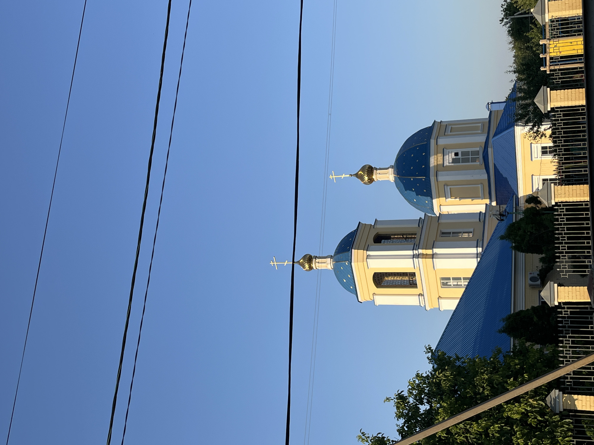 The image shows an exterior view of the Church of the Assumption of the Blessed Virgin Mary in Kotovo, Russia. The church features a yellow facade with white trim and a blue roof. Two blue domes topped with golden crosses are visible, one on a bell tower and another on the main body of the church. The sky is clear and blue, with two power lines crossing the frame horizontally. In the foreground, a metal fence with brick posts separates the viewer from the church. Trees and greenery surround the building, suggesting a suburban or rural setting. No people are present in the image.