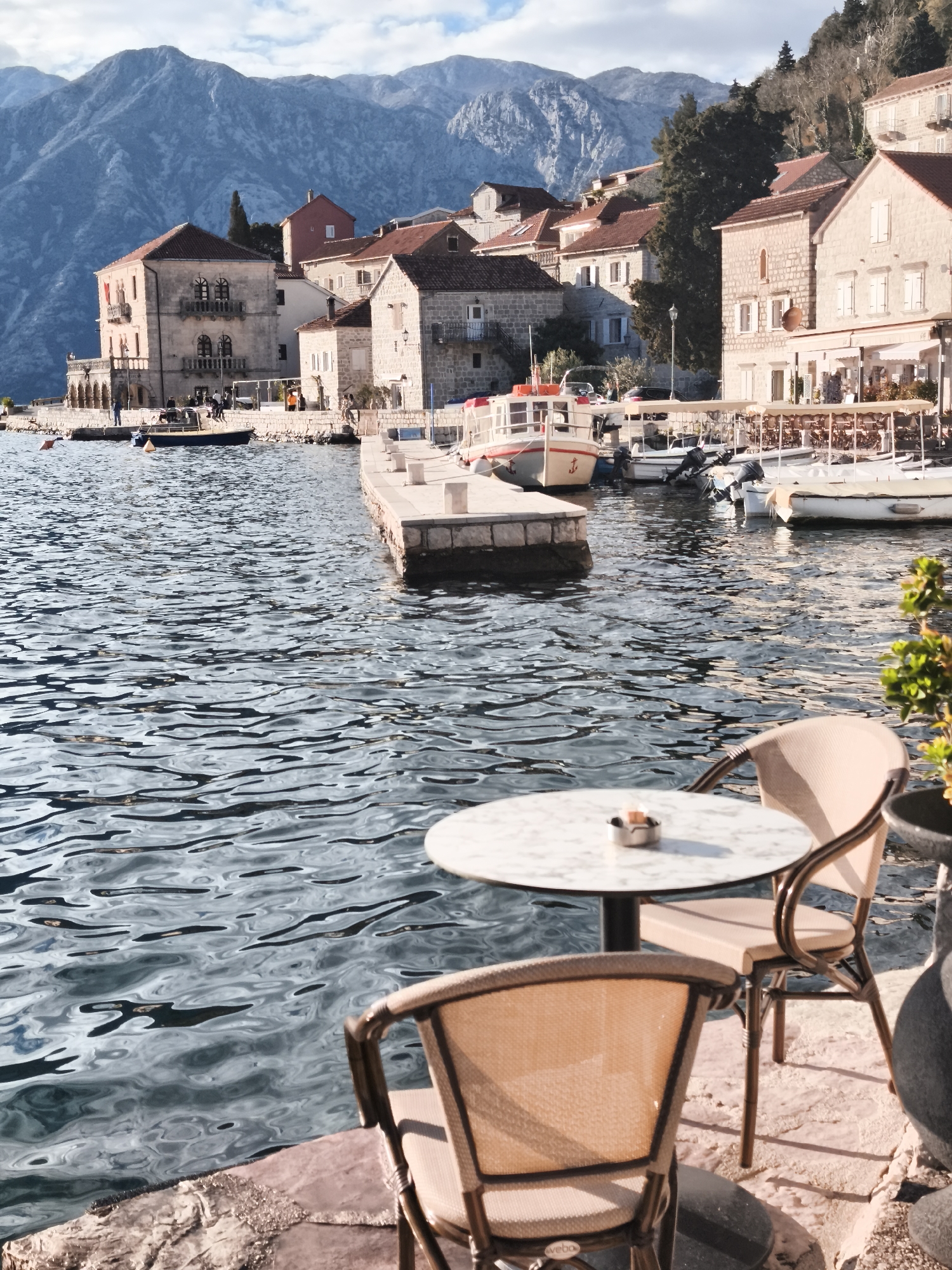 The image shows a waterfront scene in Perast, Montenegro. In the foreground, two rattan chairs and a small, round marble table are arranged on a stone patio. A metal ashtray sits on the table. Beyond the patio, a body of water leads towards a stone pier. Several small boats are docked at the pier, and along the waterfront are buildings with red-tiled roofs, typical of the area's architecture. In the background, mountains rise into the sky under a partly cloudy sky. No people are distinct in the foreground, but there are some figures visible on the shoreline near the buildings.