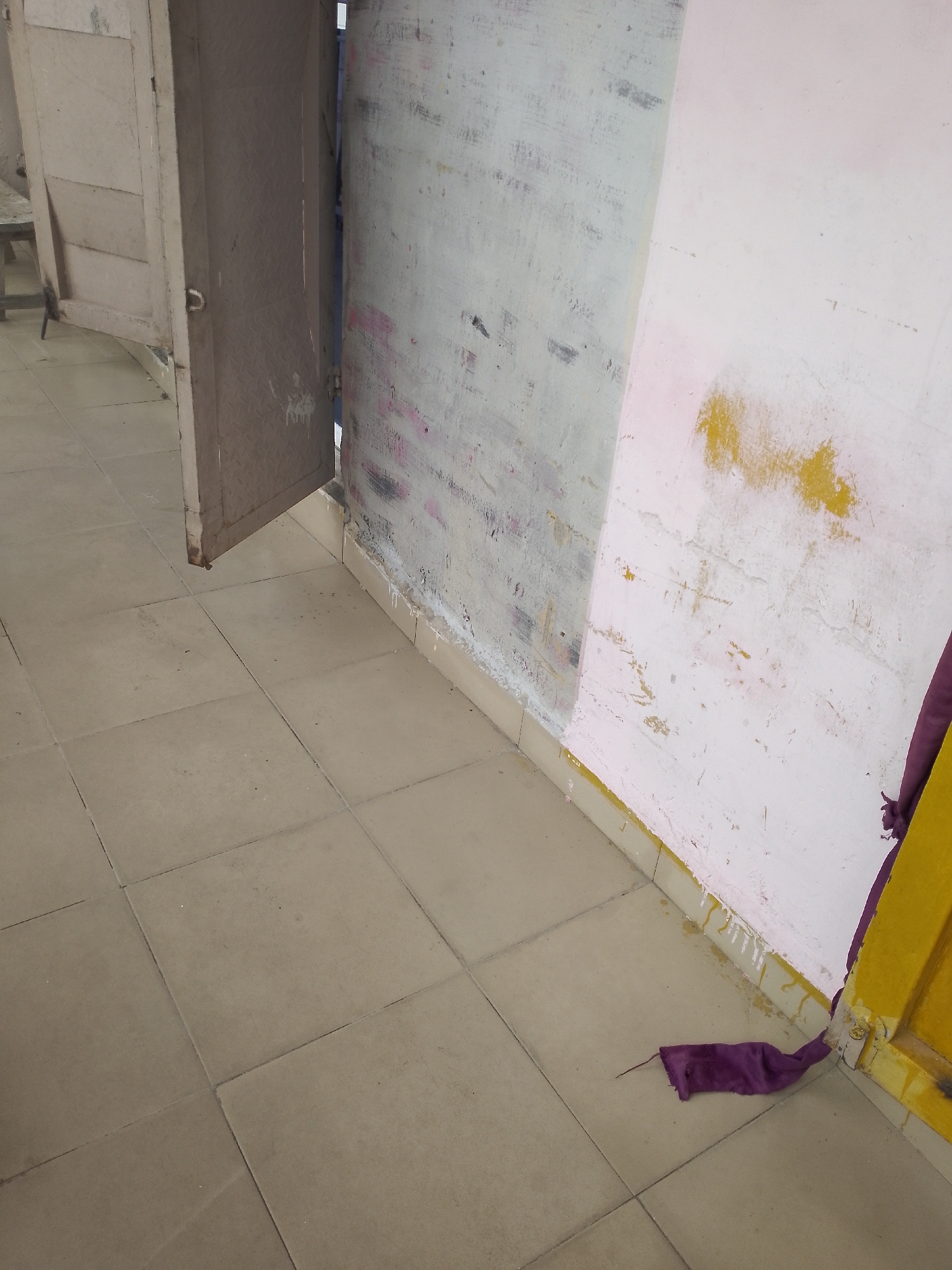 An interior view depicts a narrow hallway with beige tiled flooring. To the left, a slightly ajar, weathered wooden door is visible, revealing a glimpse of a table and chair. The hallway leads towards a corner where two walls meet. The left wall is painted a light gray with visible patches of pink and dark gray paint or stains. The right wall is predominantly pink, with significant patches of yellow and white paint, some appearing to be drips or smudges. Along the base of the walls, a cream-colored skirting board is present, with white and yellow paint drips extending onto the tiles in places. A section of a yellow doorframe is visible on the far right, partially obscured by a draped piece of dark purple fabric that trails onto the tiled floor. The scene suggests an indoor environment, possibly a workshop or storage area, in a state of disrepair or in the process of being renovated.