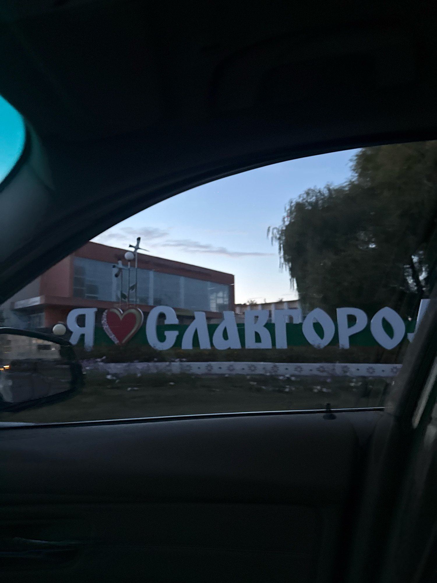 The image captures a view from inside a vehicle, looking outwards at a sign that reads "Я ♡ Славгород" which translates to "I Love Slawharad". The sign features large white letters with a red heart symbol in the center replacing the word "love". The sign is situated in front of a modern building with a facade of orange and glass. Behind the building, a tree with green foliage is visible against a pale blue sky with scattered clouds. The foreground of the image is dominated by the interior of the car, including the dark interior panels of the door and the car's ceiling. The overall scene suggests a roadside view in the city of Slawharad, Belarus.