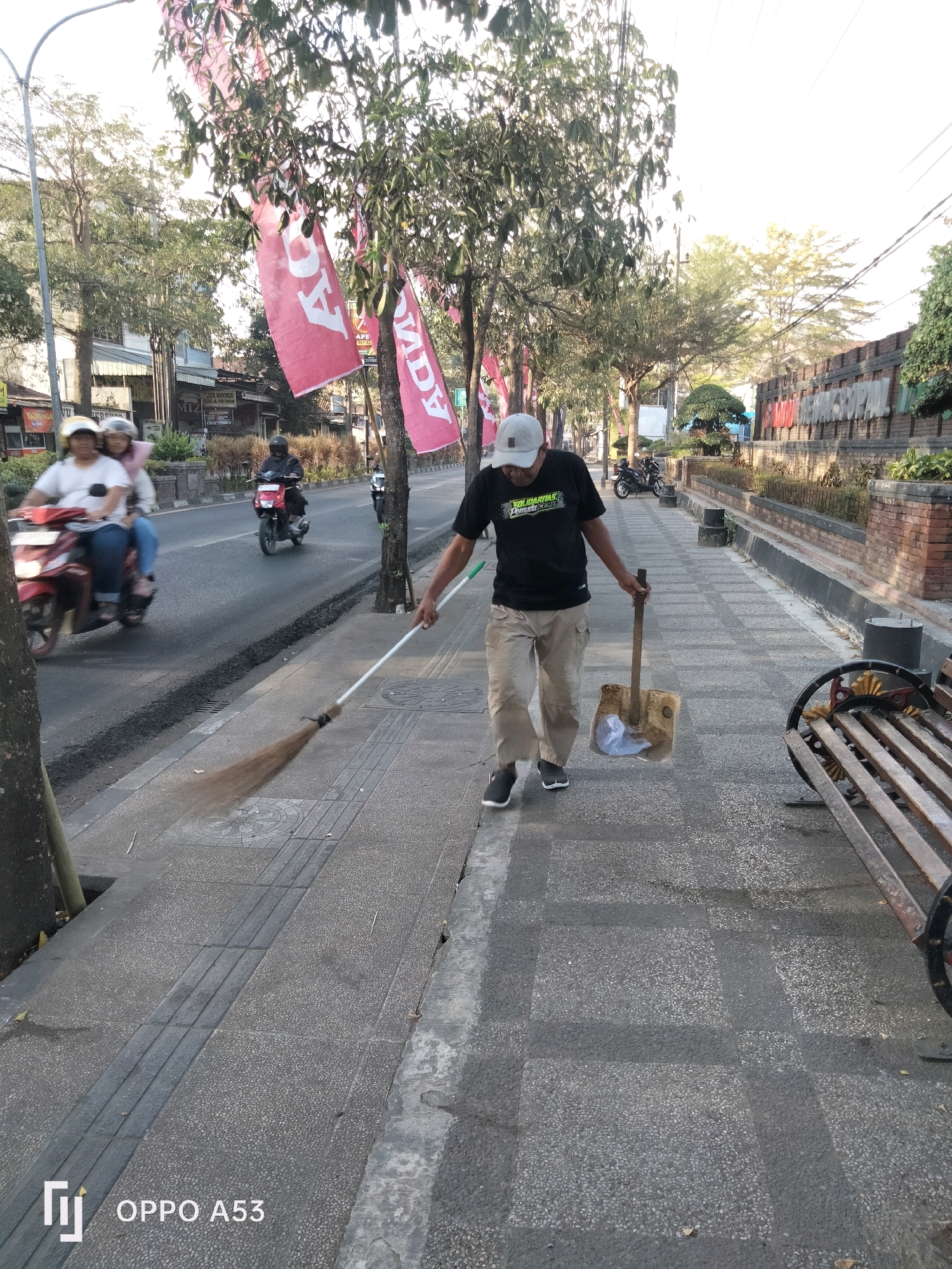 A man sweeps a sidewalk in Kelutan, Indonesia. He is dressed in a black t-shirt, tan pants, a white baseball cap, and dark sneakers. He holds a broom in his right hand and a shovel with a plastic bag in his left. The sidewalk is paved with grey stones, and a raised tactile paving strip runs along the center. To the left, a street with passing motorcycles and a few pedestrians is visible. Red banners hang from trees lining the street, and a brick wall with signage is on the right. A wooden bench is situated on the sidewalk to the right of the man. The scene is illuminated by natural daylight, casting shadows on the sidewalk.