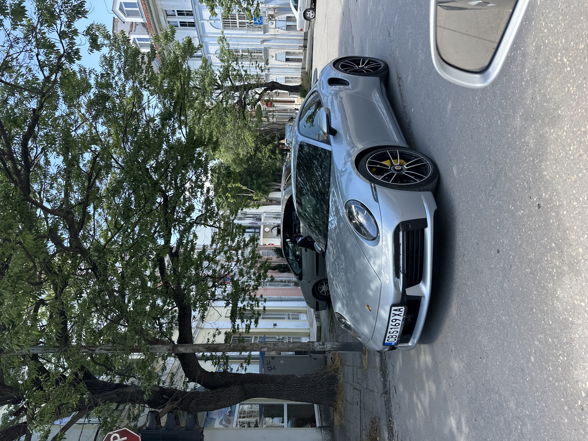 The image shows a silver Porsche 911 parked on a street in Varna, Bulgaria. The license plate reads "CB5169XA". A large tree is on the left, partially obscuring the view of buildings across the street. Another car is parked behind the Porsche. The scene appears to be a sunny day, as evidenced by the shadows and bright light. A side mirror of another car is visible on the upper right side of the picture.
