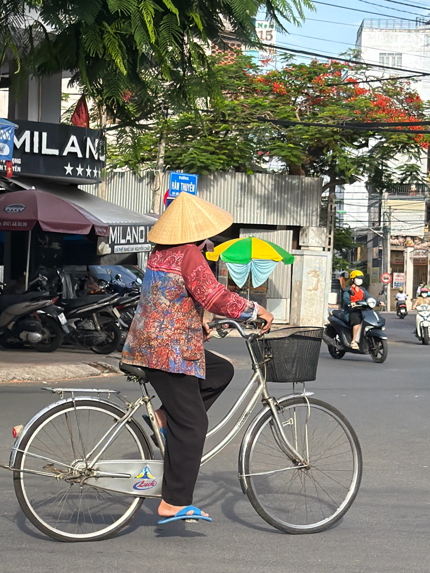The image depicts a street scene in Nha Trang, Vietnam, on a sunny day. In the foreground, a person wearing a conical hat and a colorful patterned shirt rides a silver bicycle. The bicycle has a basket on the front and a kickstand engaged. The rider is wearing black pants and blue flip-flops. Behind the bicycle, several motorcycles are parked under an awning that reads "MILANO" with stars. Further in the background, other motorcycles are on the road, and buildings line the street. A vibrant tree with red flowers is visible overhead. The scene captures a typical urban environment with traffic and local businesses.